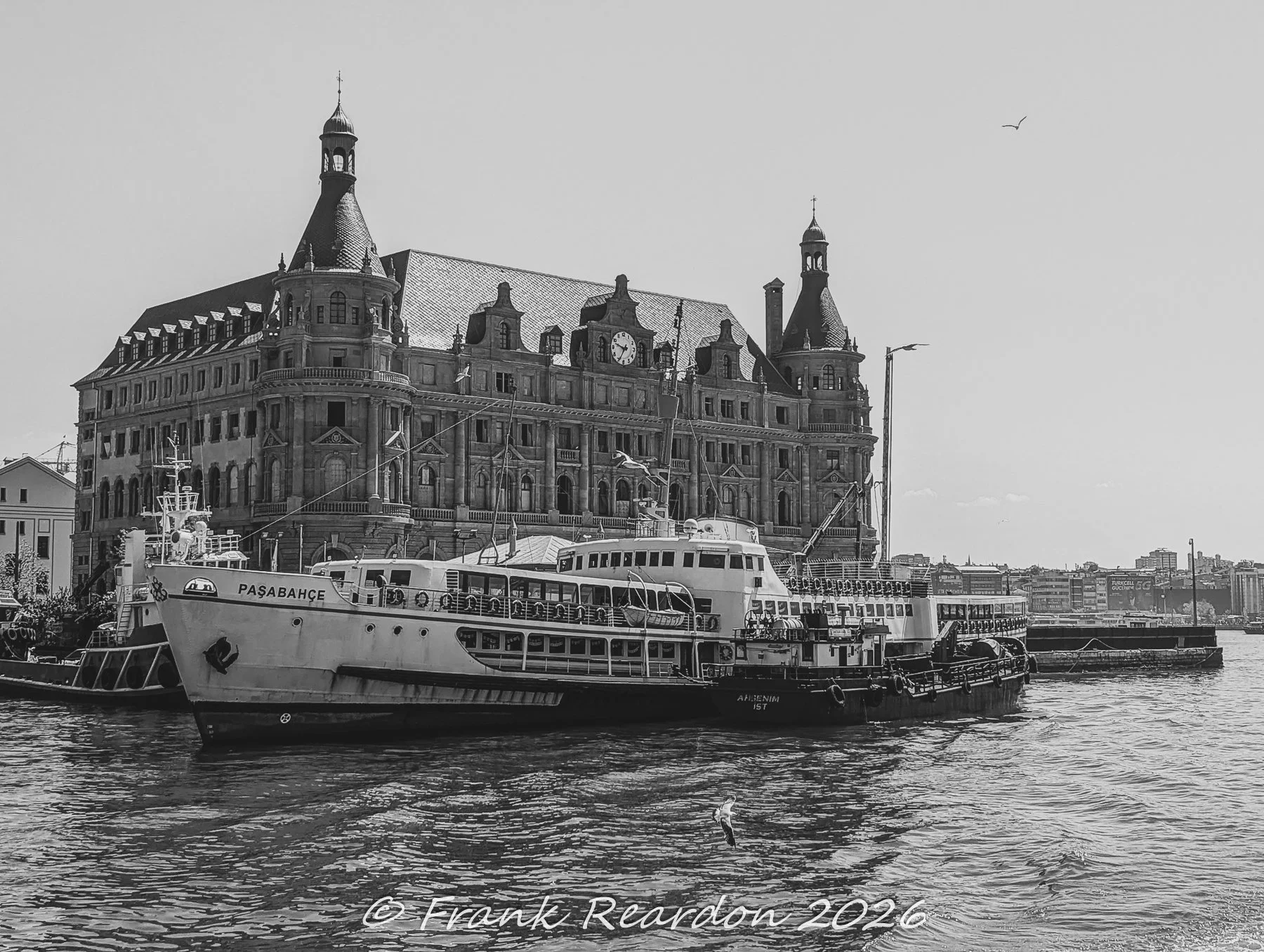 A black-and-white architectural shot of the landmark Haydarpaşa Railway Station alongside the moored Paşabahçe passenger ferry and smaller utility craft in the harbor.