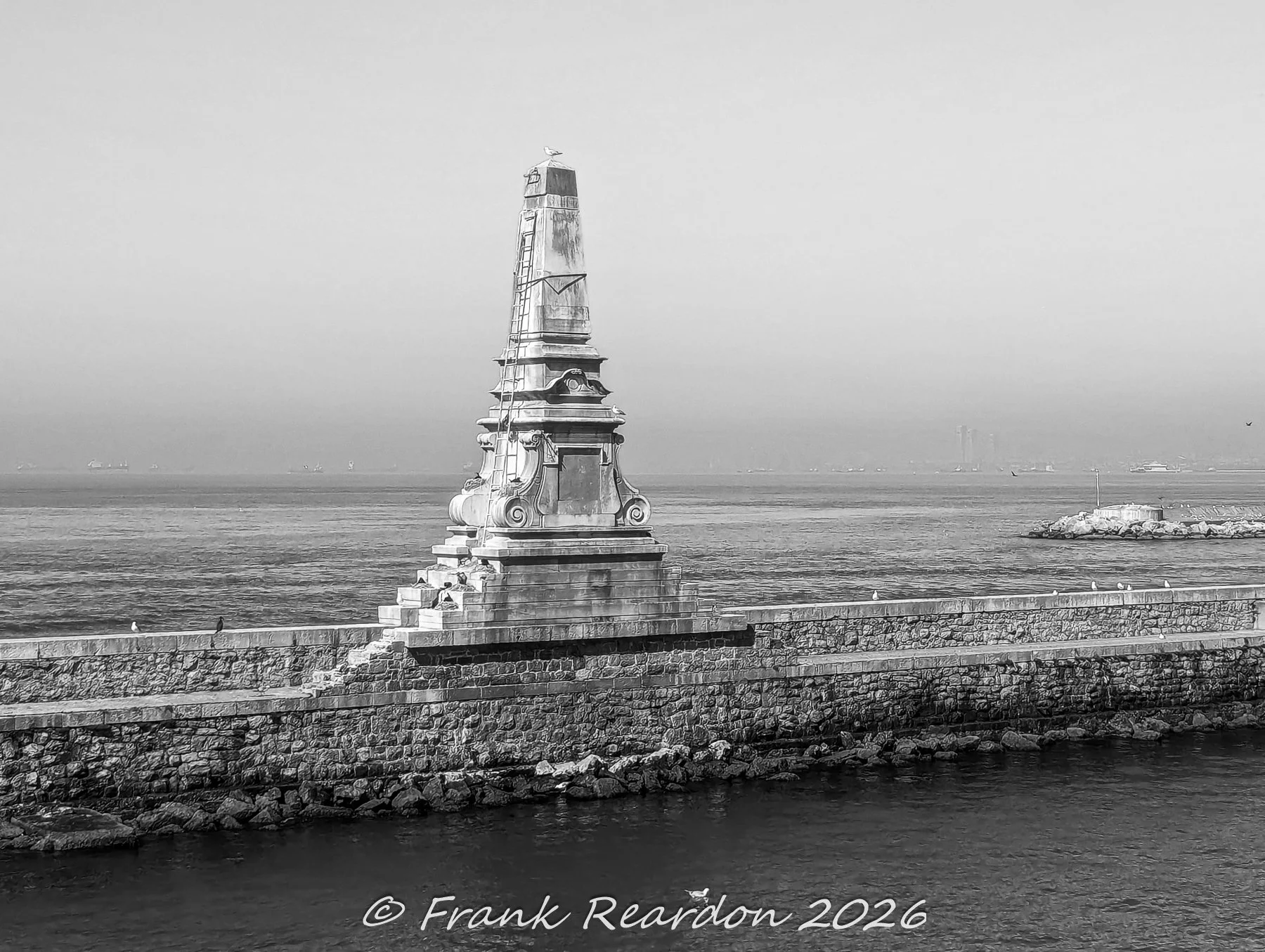 A black-and-white shot of a detailed stone obelisk and monument standing atop the long breakwater wall framing the harbor.