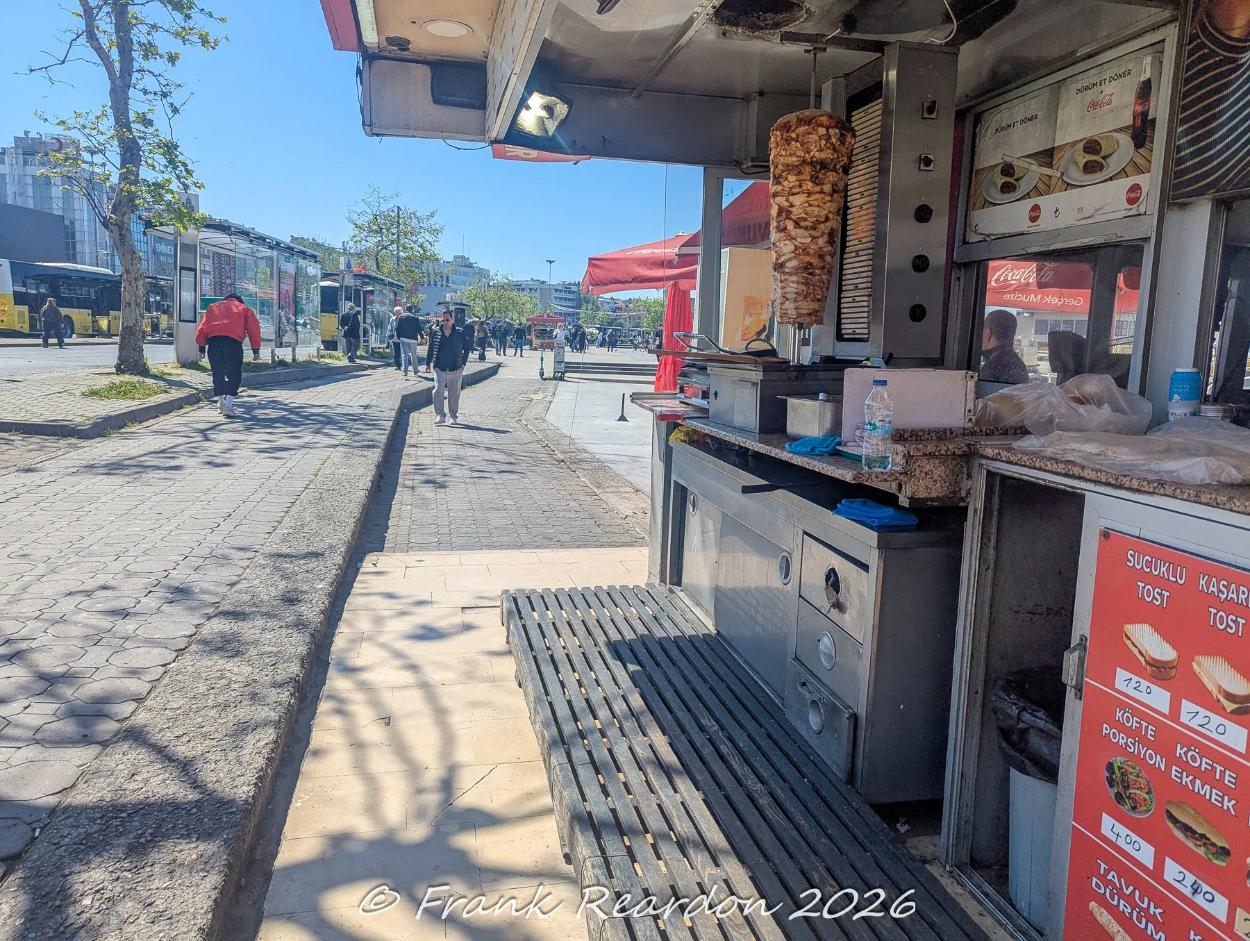 A sidewalk food kiosk featuring a tall, vertical rotisserie meat spit of döner kebab and menu signs displaying local food options like toast and dürüm.