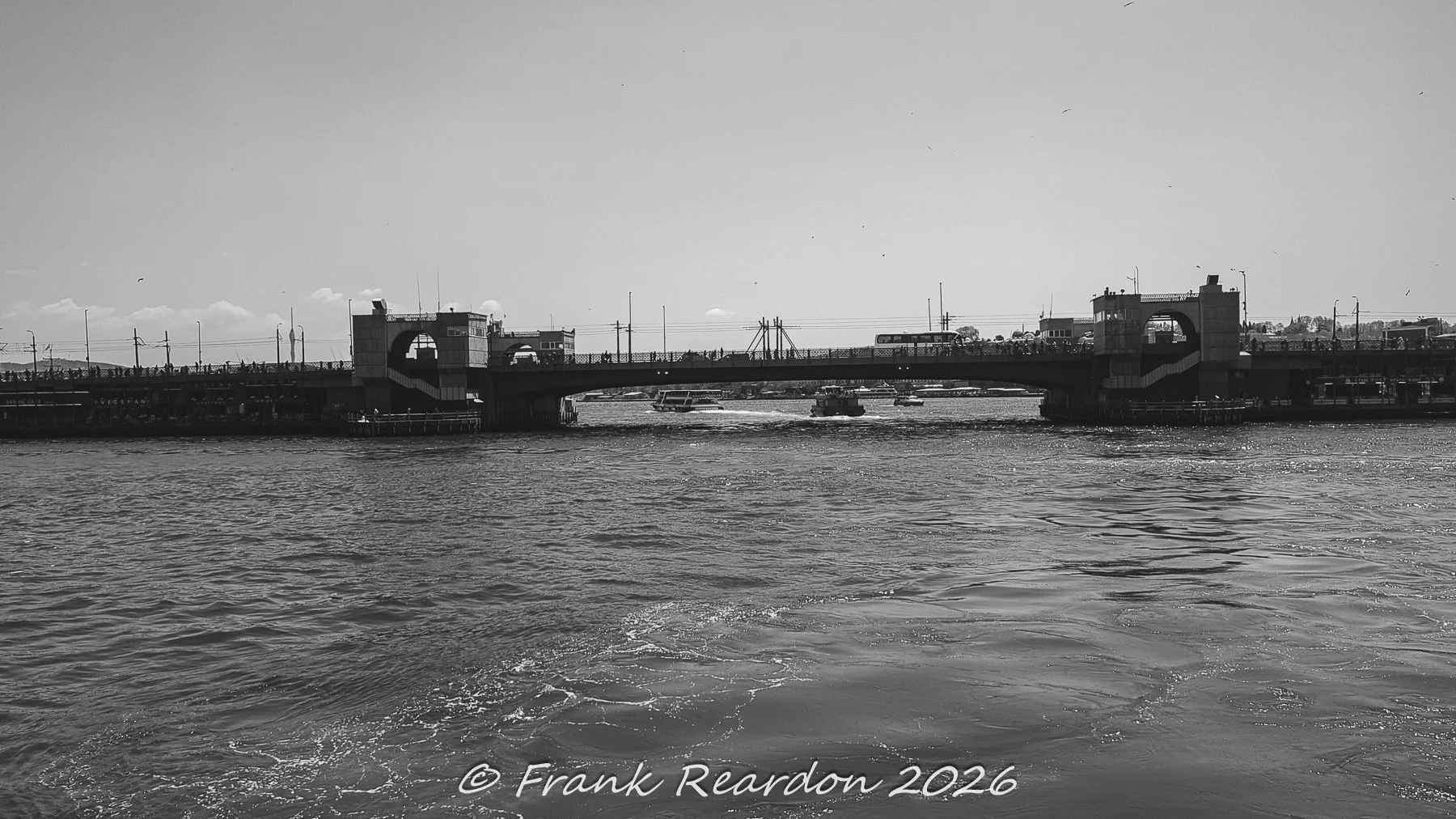 A wide black-and-white view capturing the length of the Galata Bridge with commuter traffic and pedestrians crossing against the bright daytime sky.