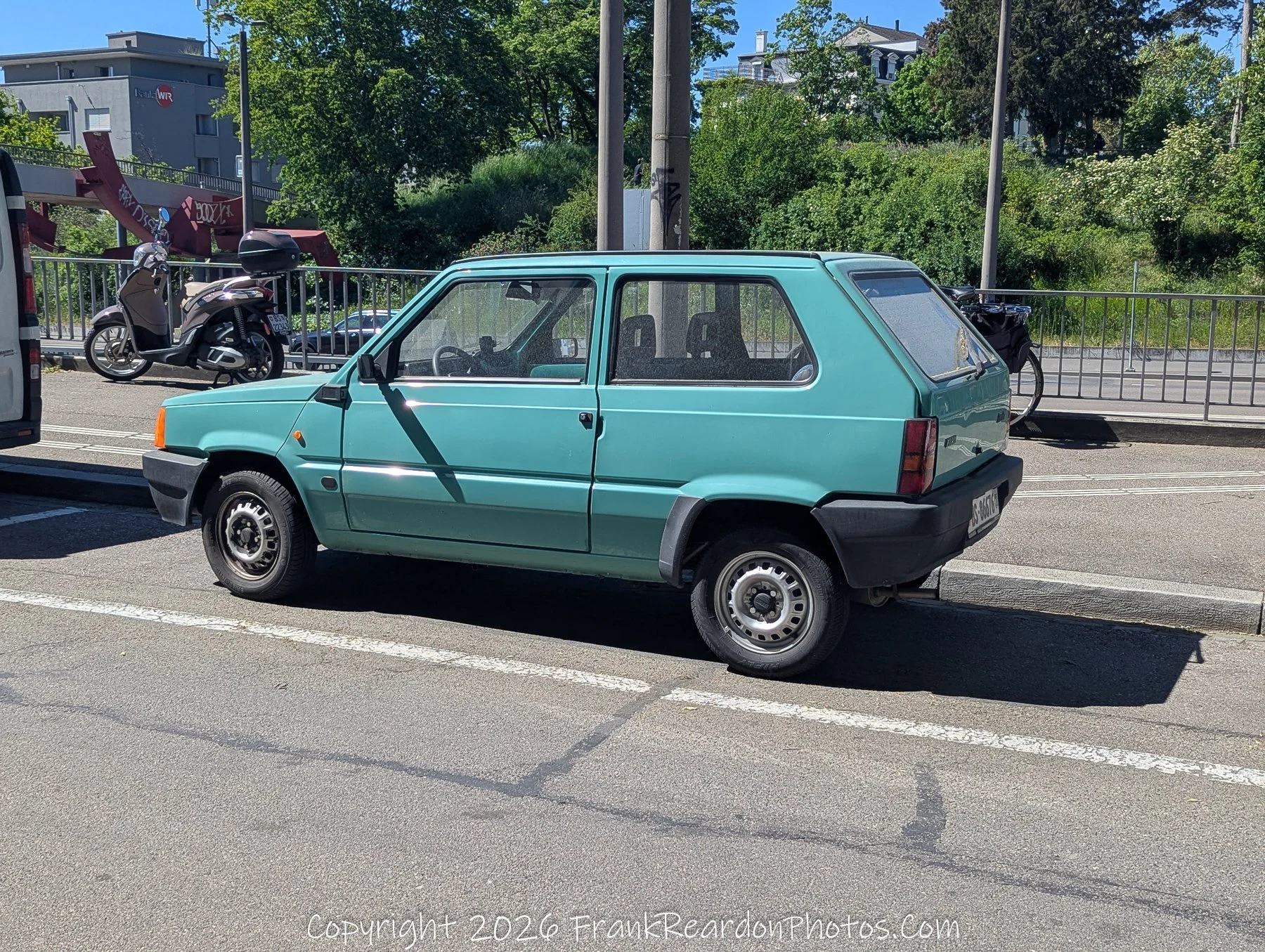 "Vintage Teal" A classic, compact teal car parked on the street, captured in the clear light of day with the red sculpture of Parkhaus Steinen visible in the background.