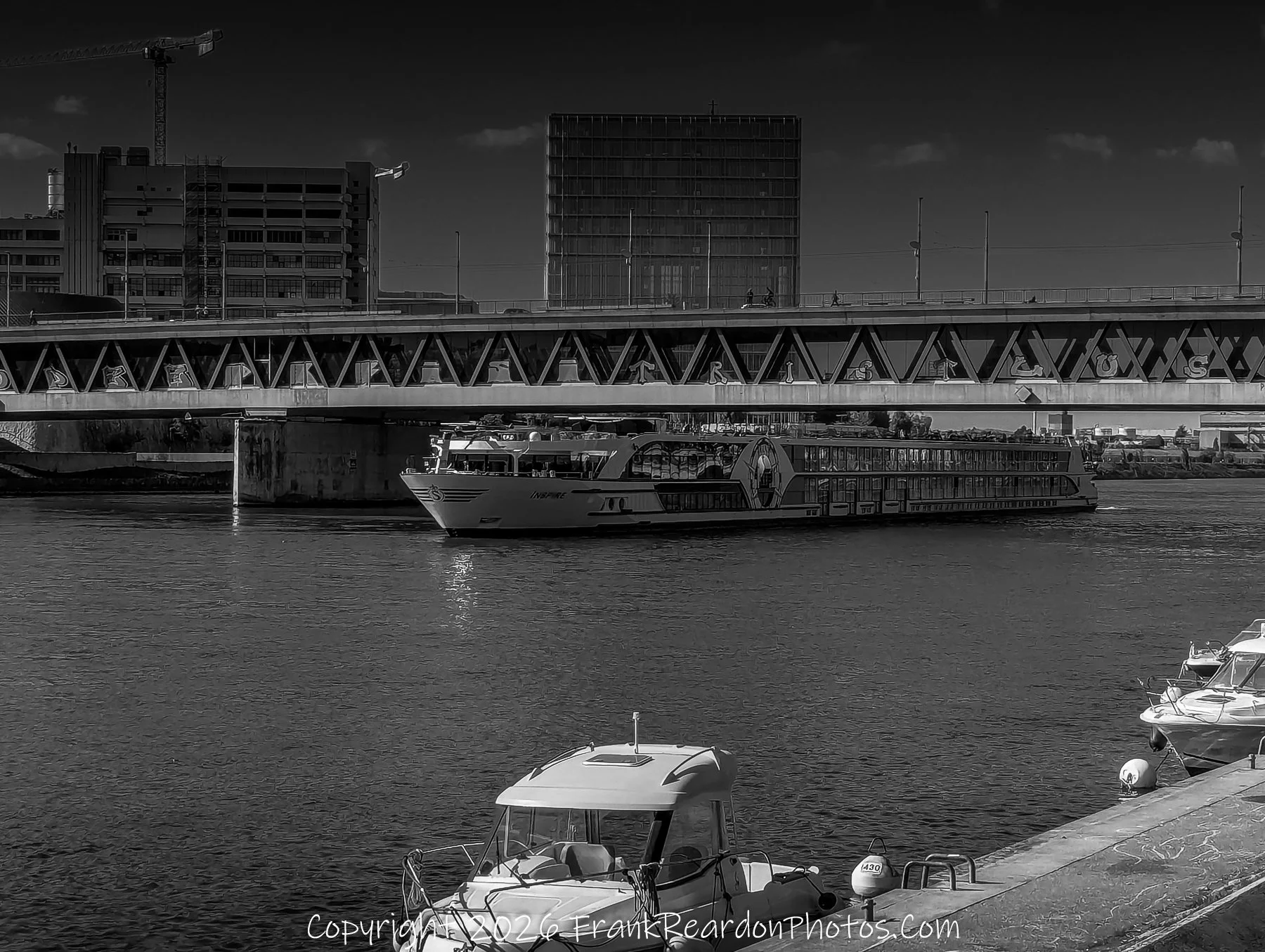 "River Transit and Docked Boats"
A black-and-white scene capturing a large river cruise ship passing under a bridge, with smaller pleasure boats docked in the foreground.
