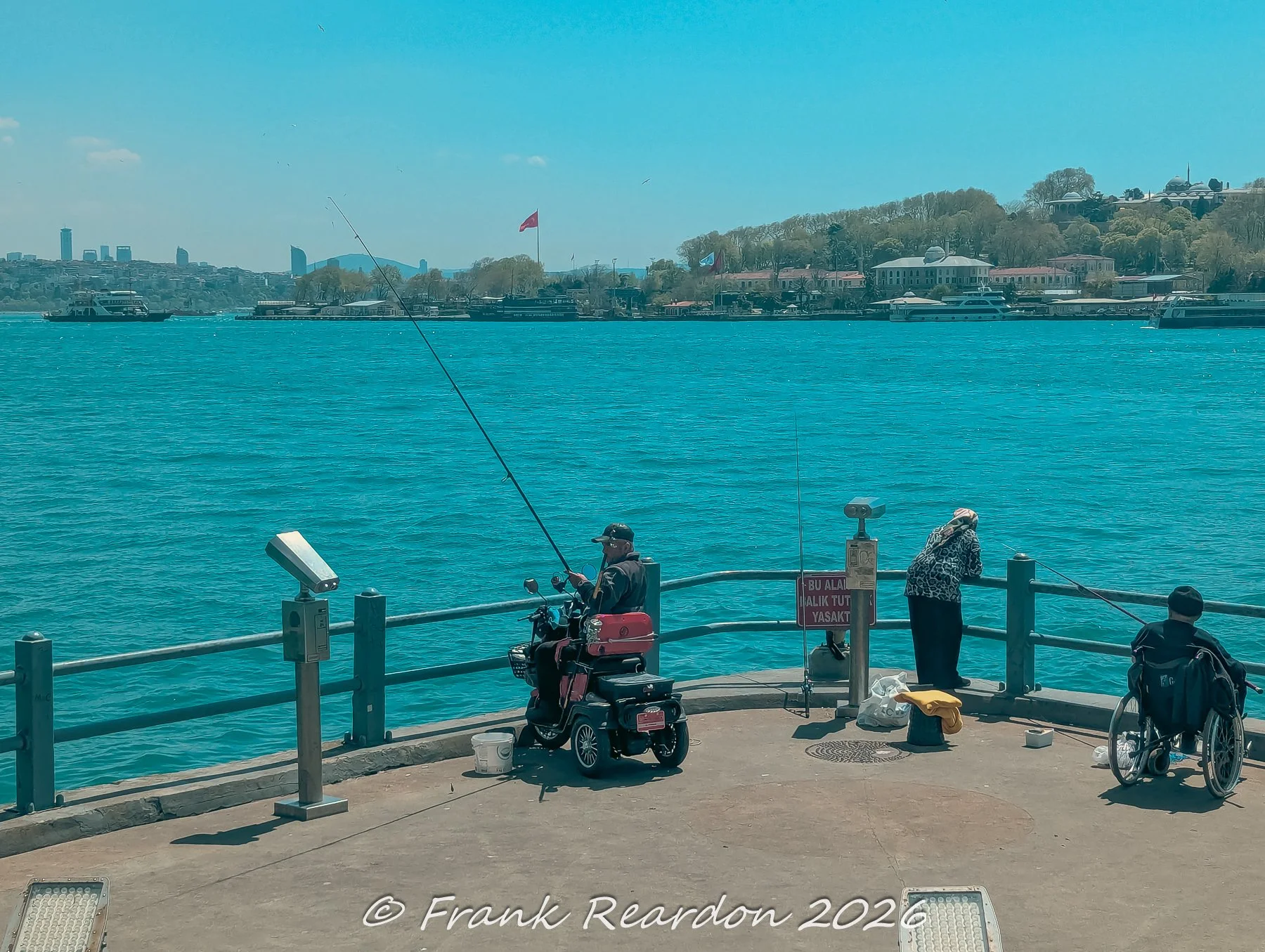 A scenic daytime shot of locals fishing along the waterfront promenade, featuring an angler seated on a mobility scooter alongside others enjoying the view of the Bosphorus.