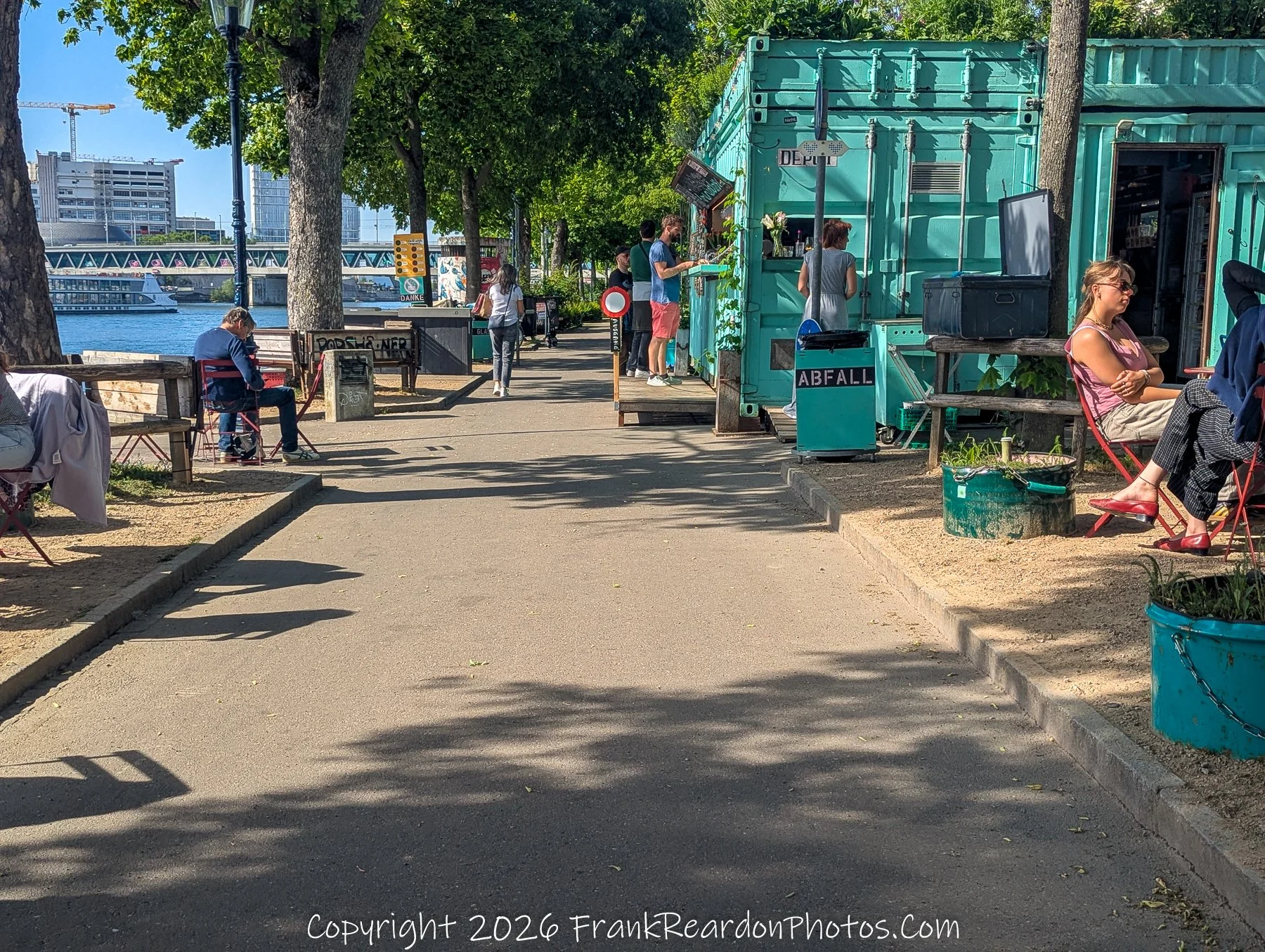"Afternoon Socializing by the River Depot" People gathering and relaxing on a pathway next to a bright teal shipping container transformed into a kiosk or café.