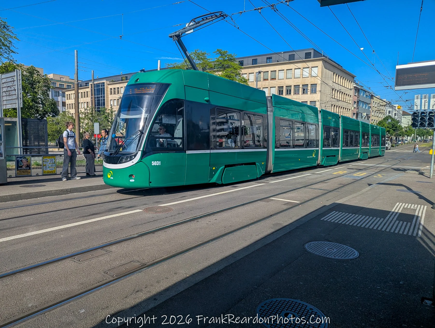 "Modern Transit at the Station" A vibrant green public transport tram arriving at a stop, with passengers waiting on the platform under a clear blue sky.