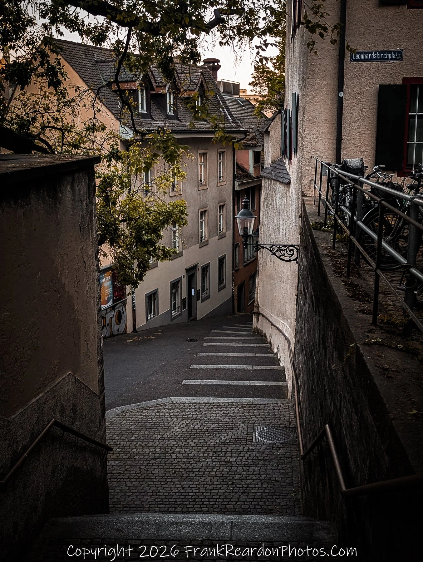 "Urban Descent" A set of stairs descending from a street corner area with buildings and a street lamp.