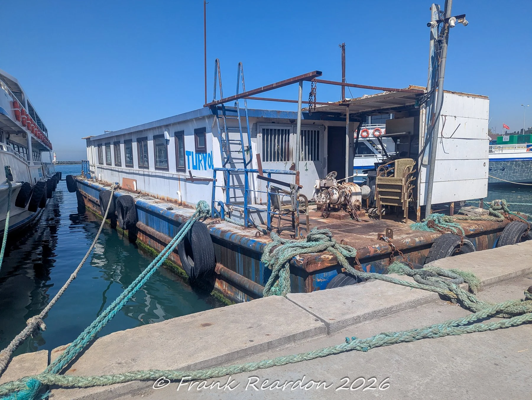 A weathered, blue-and-white service boat labeled Turyol tied to a concrete pier with thick green ropes, featuring old metal stairs, a small cabin, and a deck full of clutter.