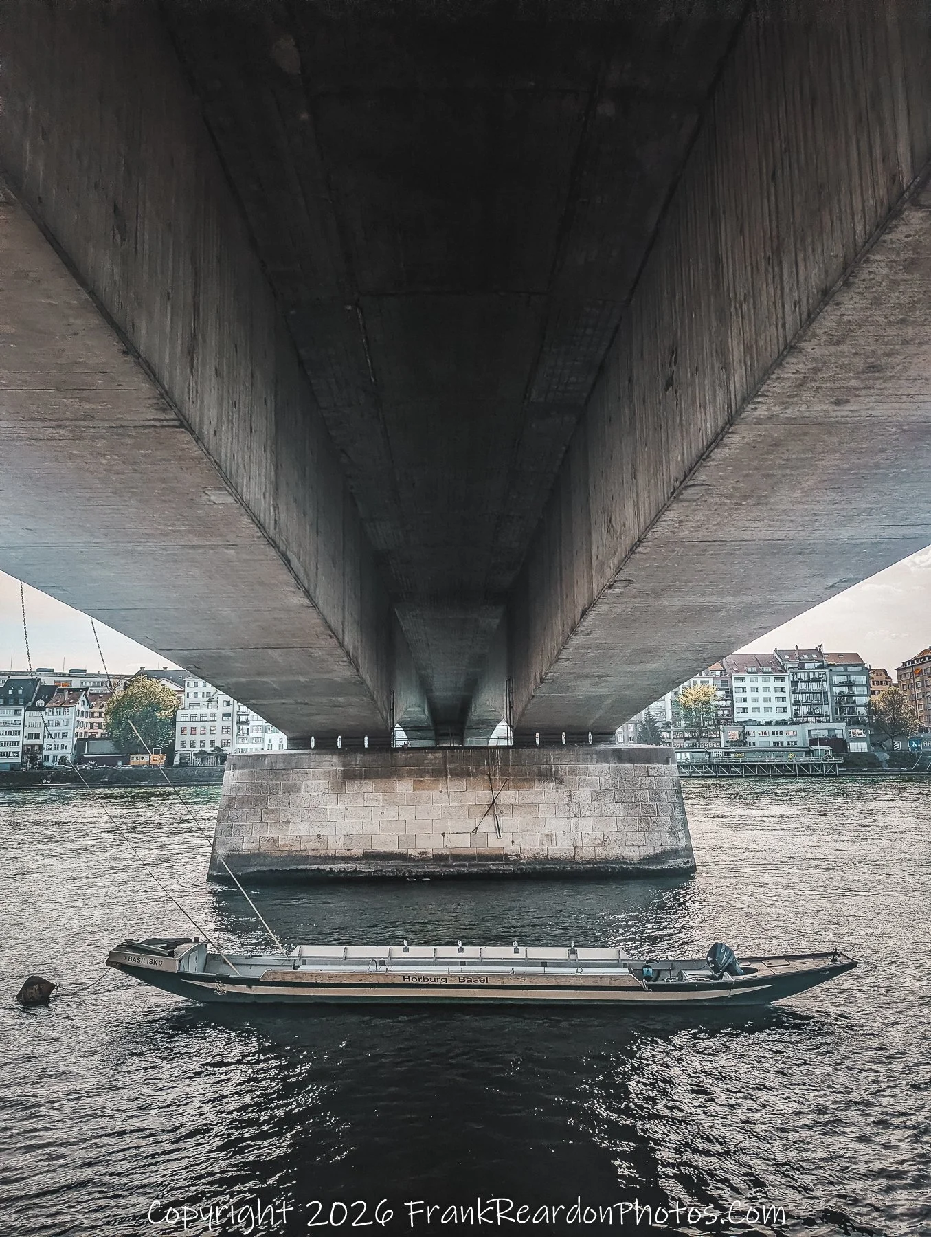 "Tranquil Waters Under the Concrete Span" A view looking beneath a massive concrete bridge structure at the river, where a single boat named "Horburg Basel" is anchored.