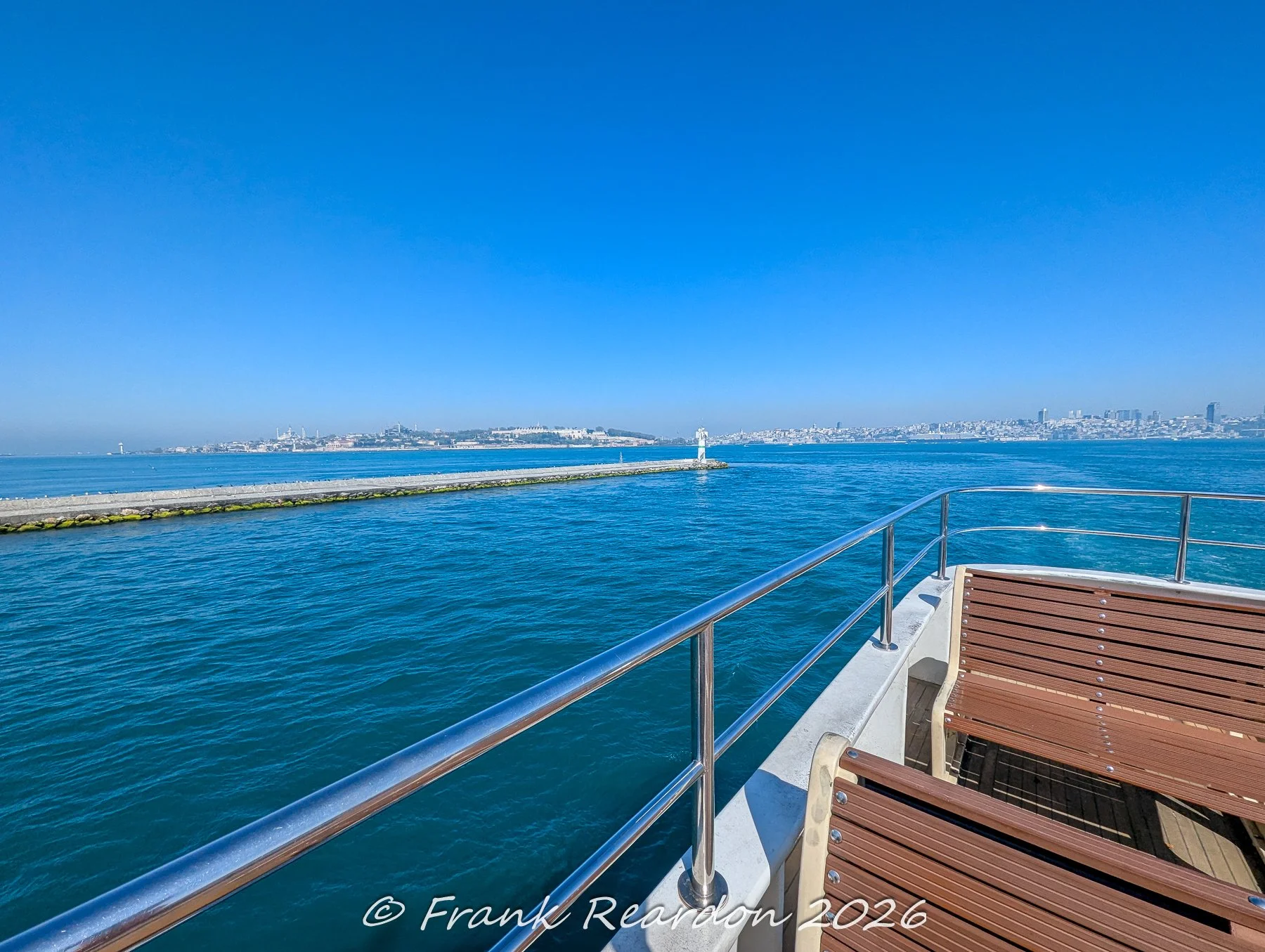 Looking past the wooden seating and steel railings on the bow of a passenger ferry toward a lighthouse marking the harbor entrance in Istanbul.