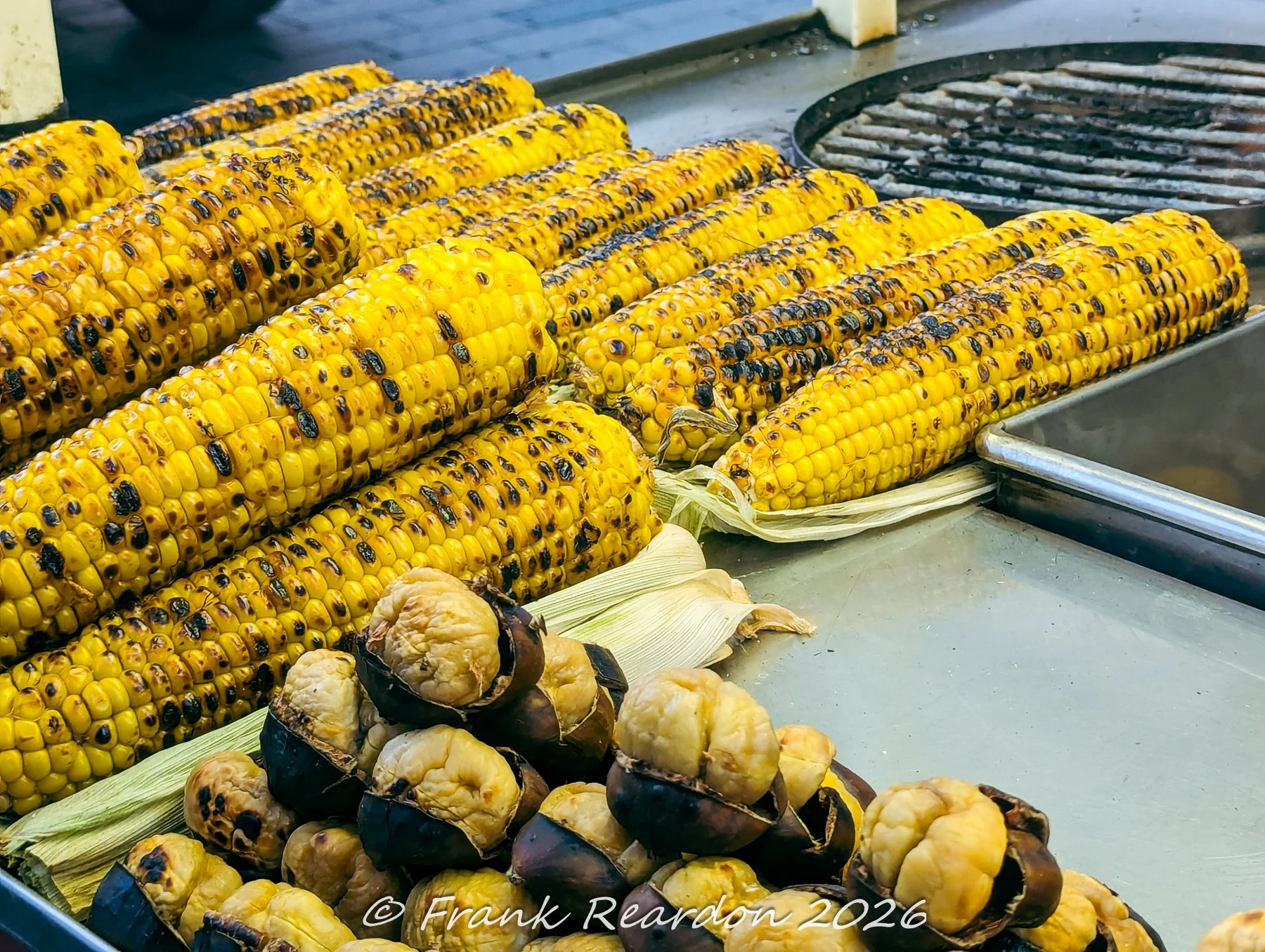 A close-up shot of golden grilled corn on the cob alongside open roasted chestnuts on a vendor's cart.