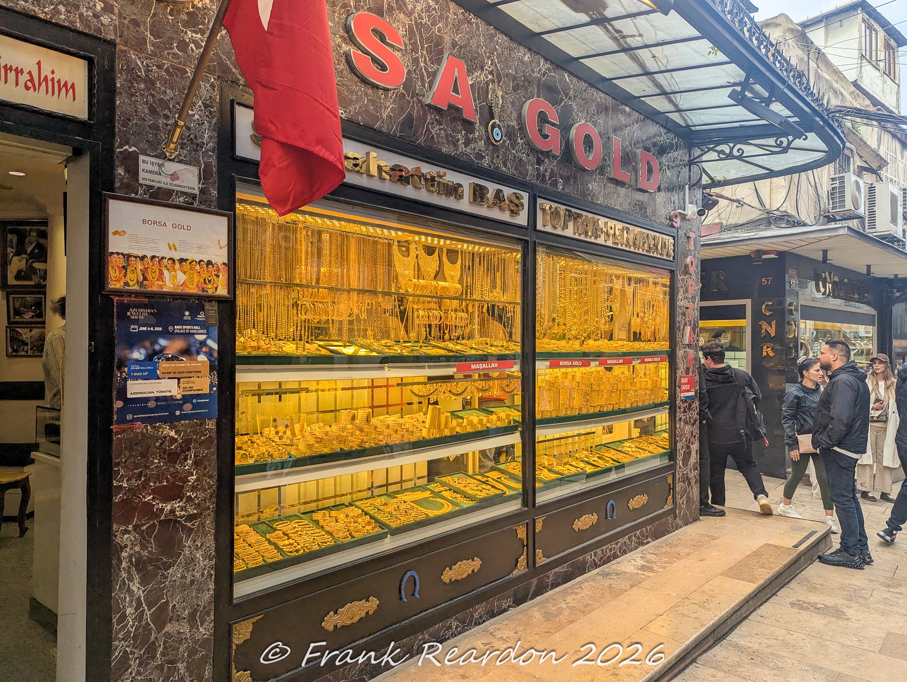 A brightly lit storefront window packed from top to bottom with cascading gold necklaces, bracelets, and ornaments along a commercial street adjacent to the Grand Bazaar.