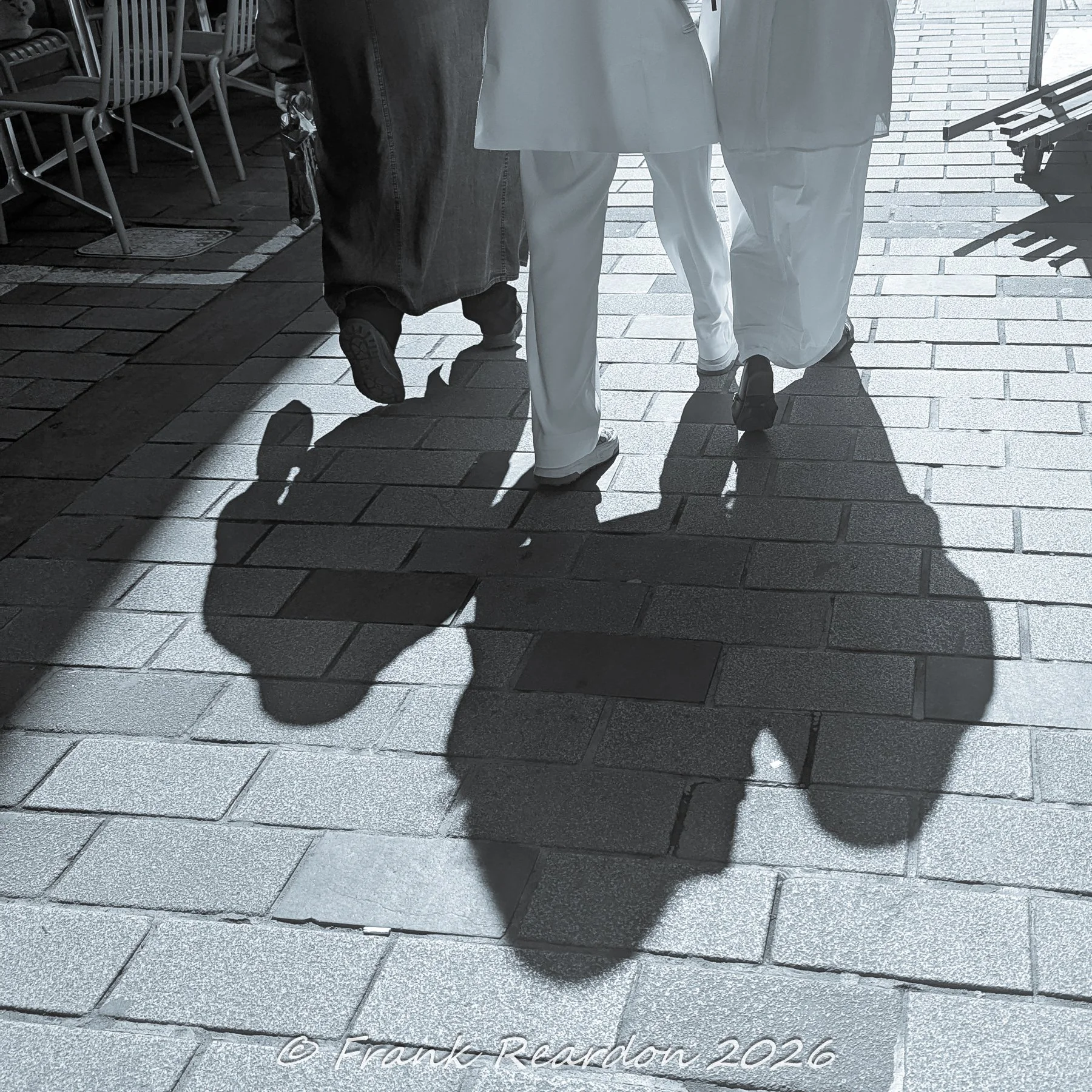 A monochrome street-level shot capturing the distinct silhouettes and shadows of pedestrians walking along the historic stone-paved sidewalks.