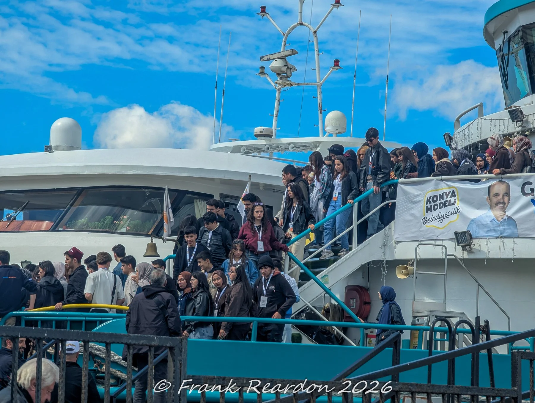 A crowd of travelers and students walking down the gangway of a passenger ferry at an Istanbul terminal, with a campaign banner visible on the vessel.