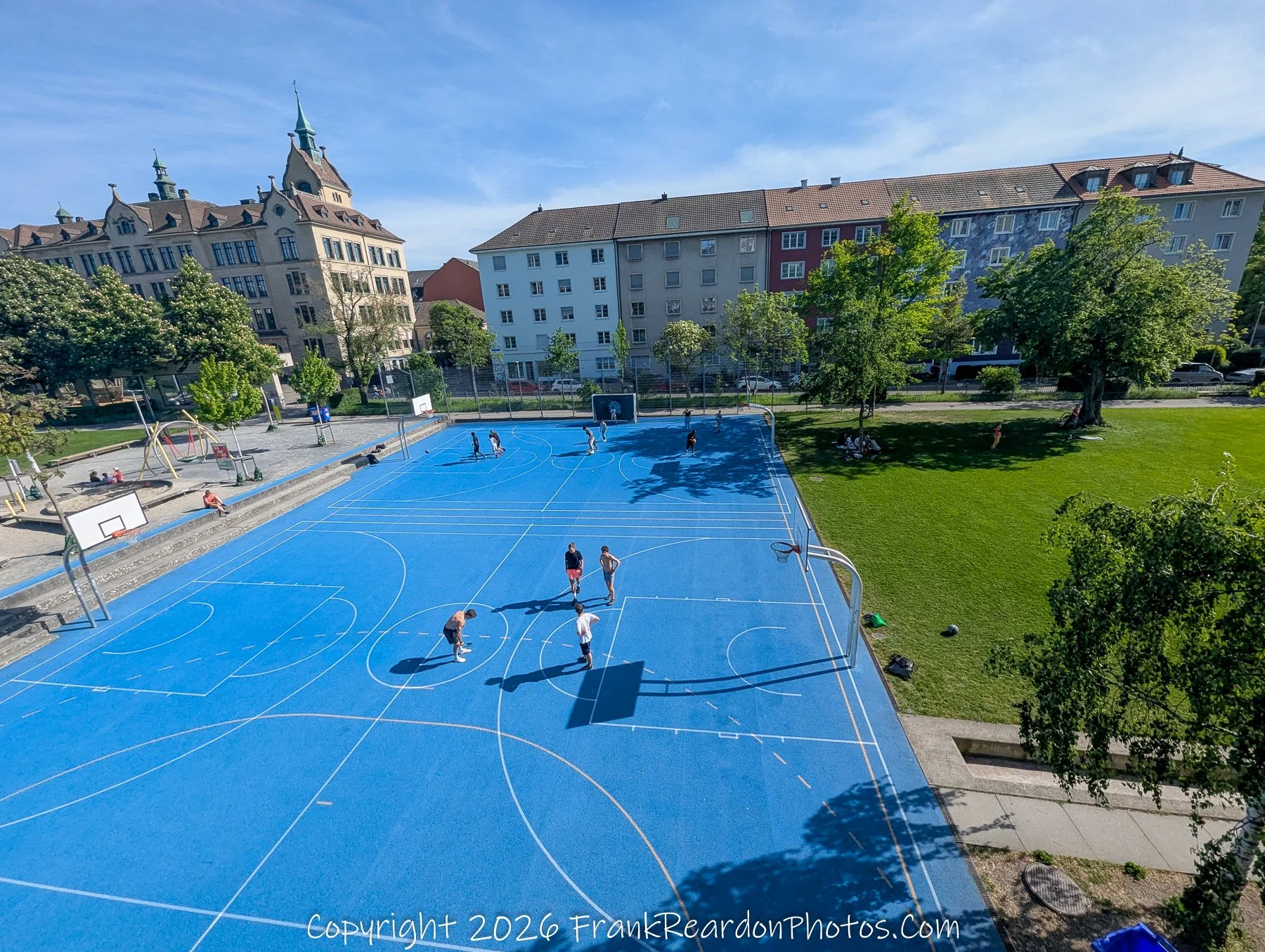 "Sunny Afternoon on the Blue Court" An elevated view of a vibrant blue outdoor sports court where a group of people are gathered playing basketball near a grassy park area.