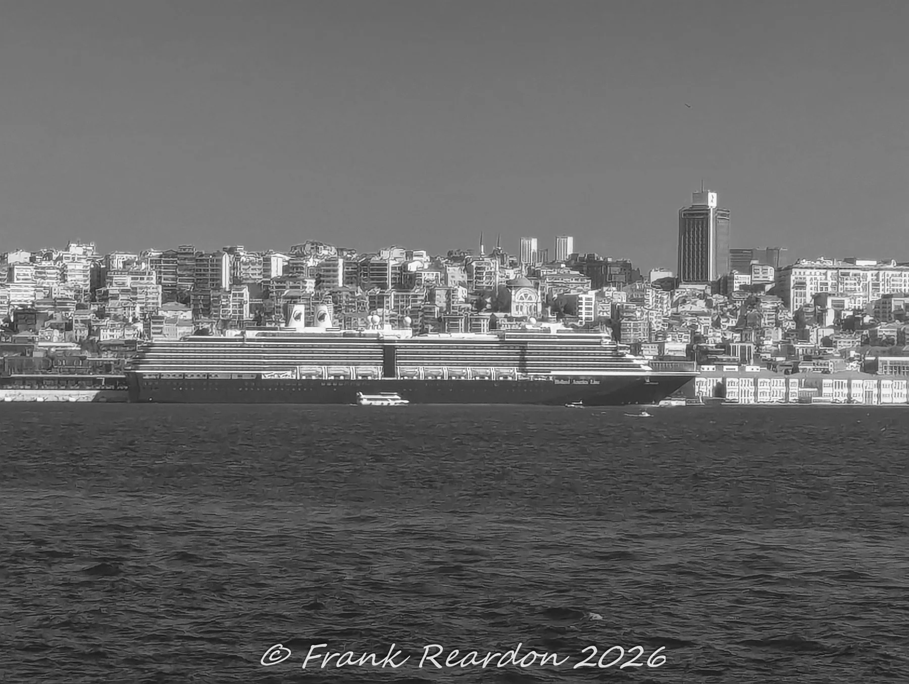 A monochrome view of a large cruise liner moored along the waterfront, with the hillside residential blocks of Istanbul rising in the background.