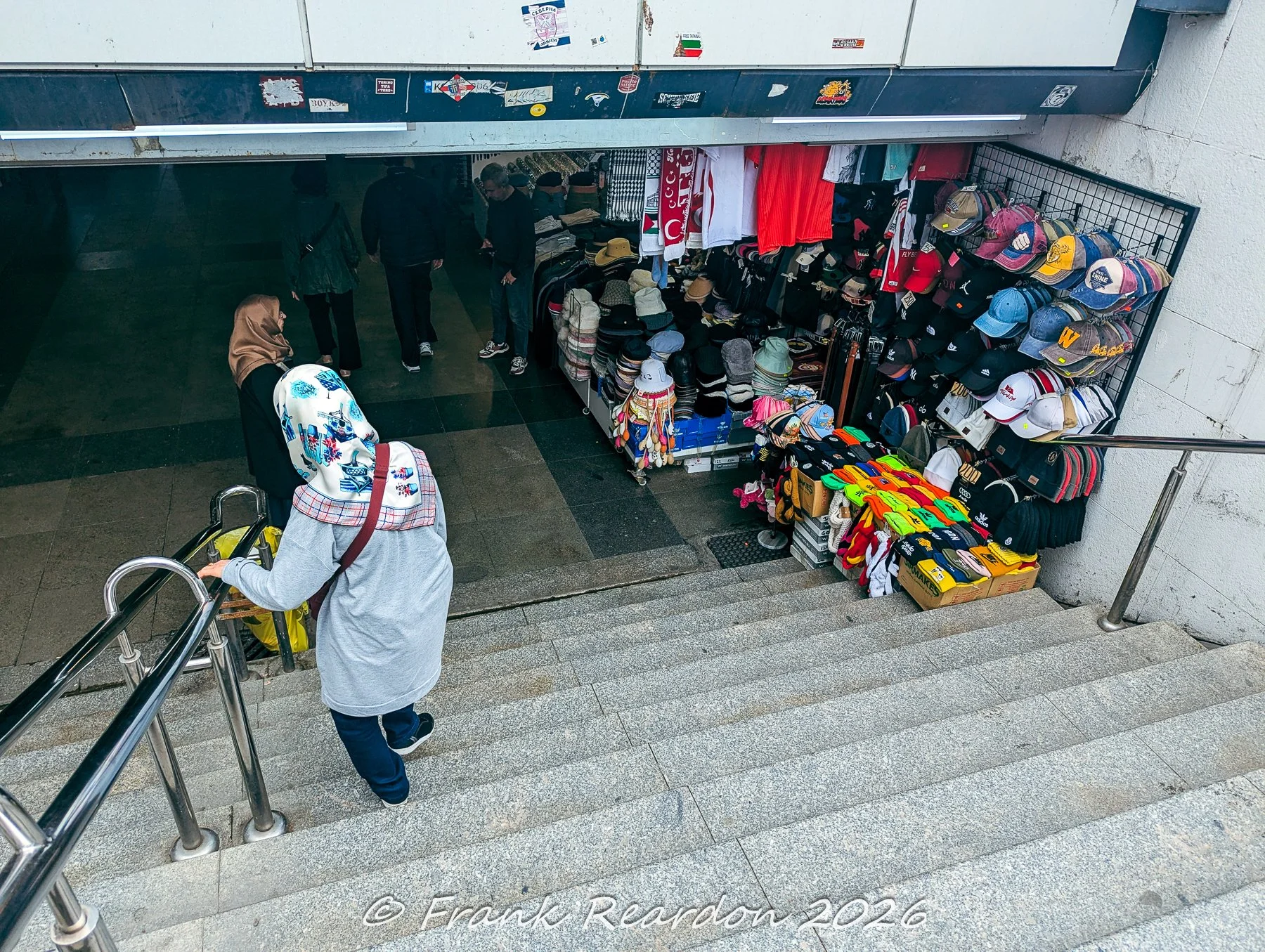 Pedestrians walking down concrete steps into an underground pedestrian underpass featuring an outdoor clothing and hat shop display.