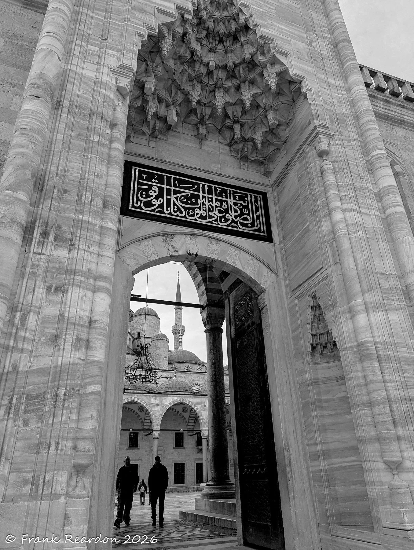 A detailed monochrome view of an intricate stone archway featuring traditional Islamic calligraphy and stone-carved patterns.