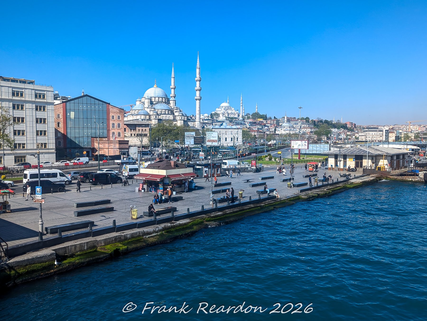 A view from the water looking across the bustling Eminönü waterfront square, featuring the prominent domes and minarets of the historic New Mosque (Yeni Cami) against a clear blue sky.