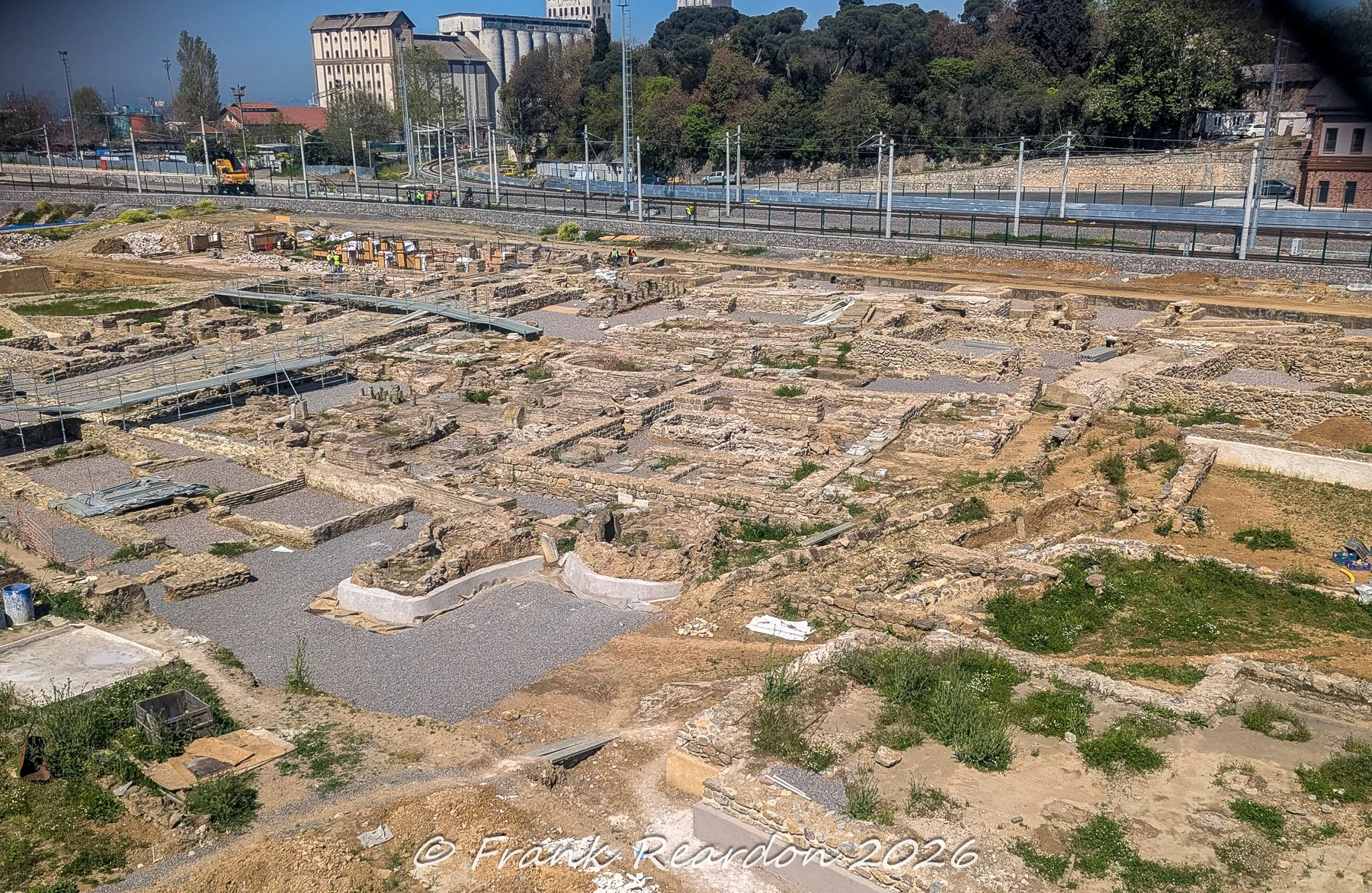 A wide-angle, elevated view of an extensive archaeological excavation site showcasing old stone foundation walls and trenches near a railway line and industrial buildings.