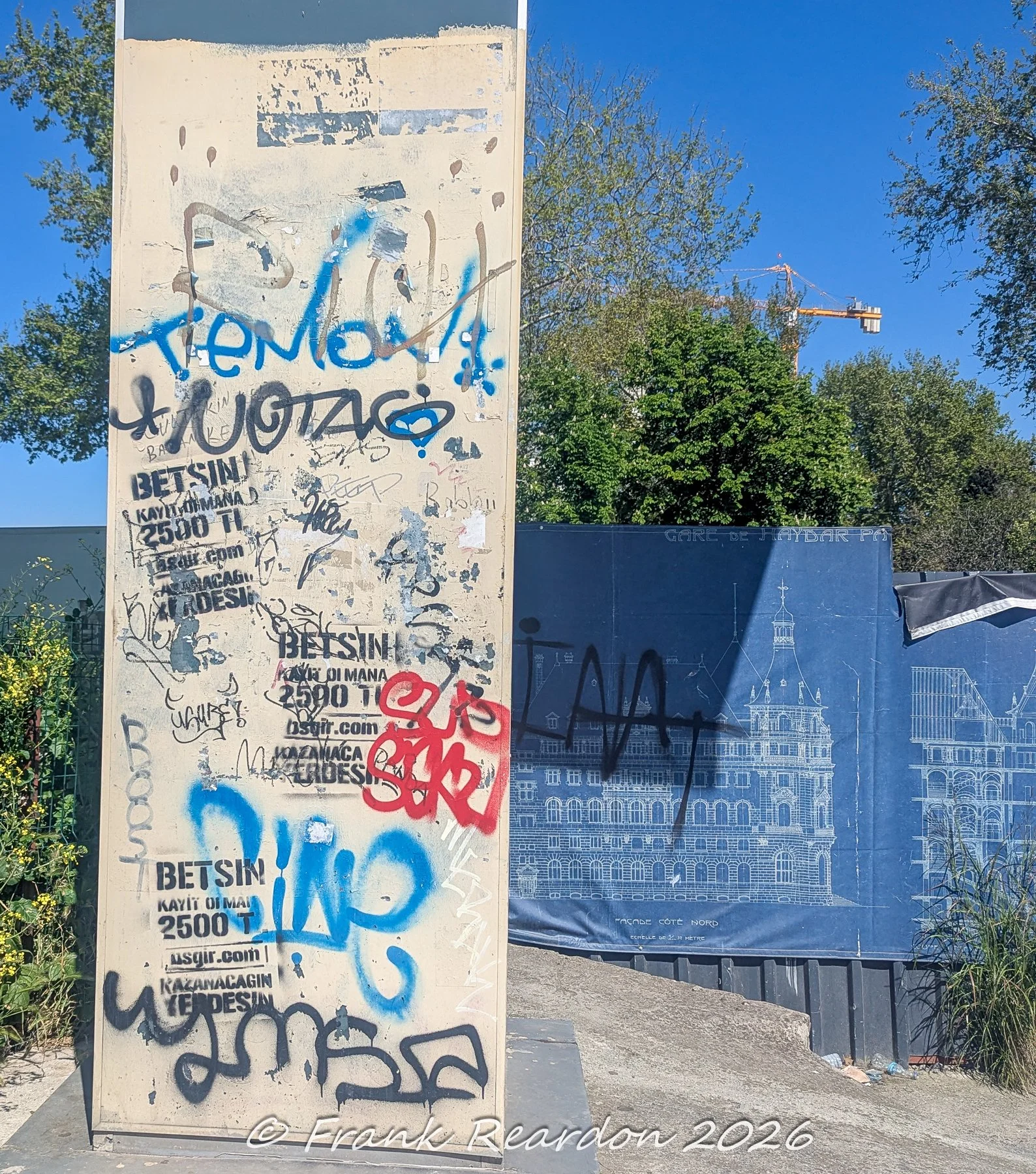 An urban concrete pillar covered in colorful graffiti tags and flyer remnants, standing next to a blue construction barrier displaying architectural blueprints for Haydarpaşa Garı.