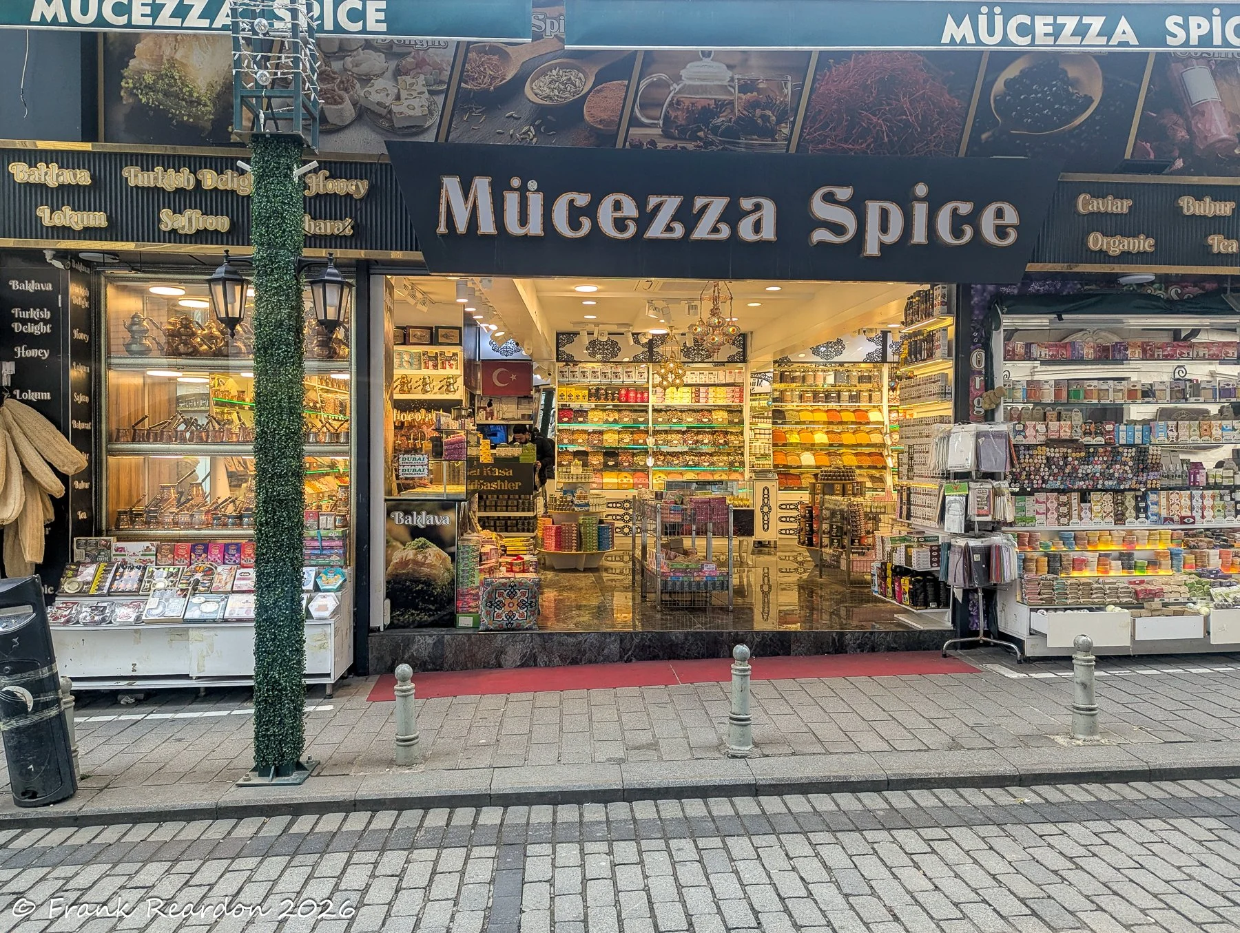 A vibrant, well-stocked spice and Turkish delight shop, "Mücezza Spice," viewed from across a cobblestone street.