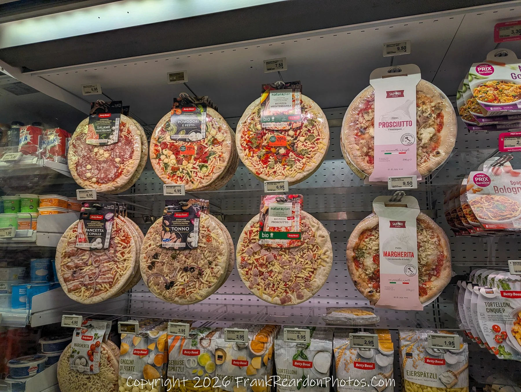 "A Selection of Basel Market Pizzas" A refrigerated grocery display case featuring various pre-made pizzas, including prosciutto, margherita, and vegetable varieties, along with other pasta products like gnocchi and spätzli.