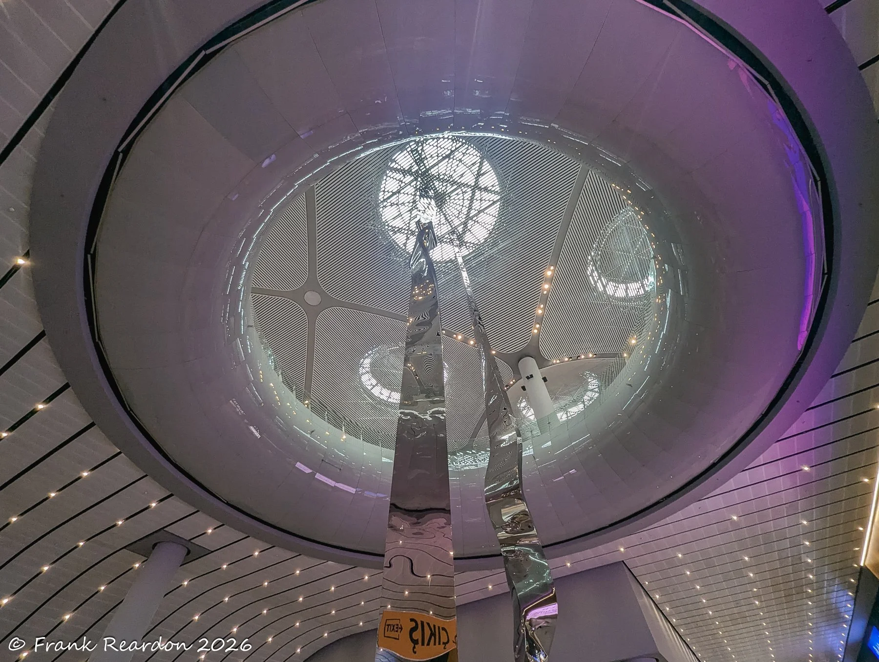 Looking up into the striking circular glass skylight and modern structural design of the Istanbul Airport interior, complemented by reflective sculptural elements.