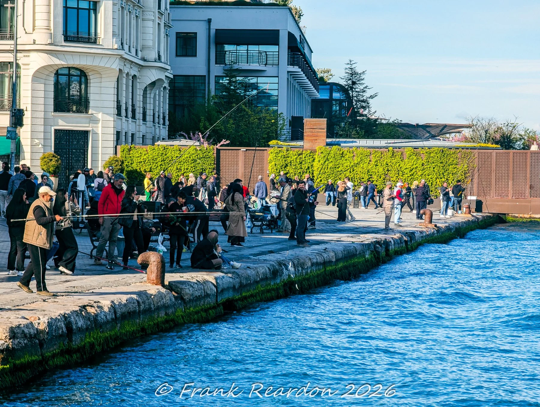 A lively scene of local residents and fishermen lining the stone promenade along the Bosphorus shoreline, casting their rods near historic architecture.