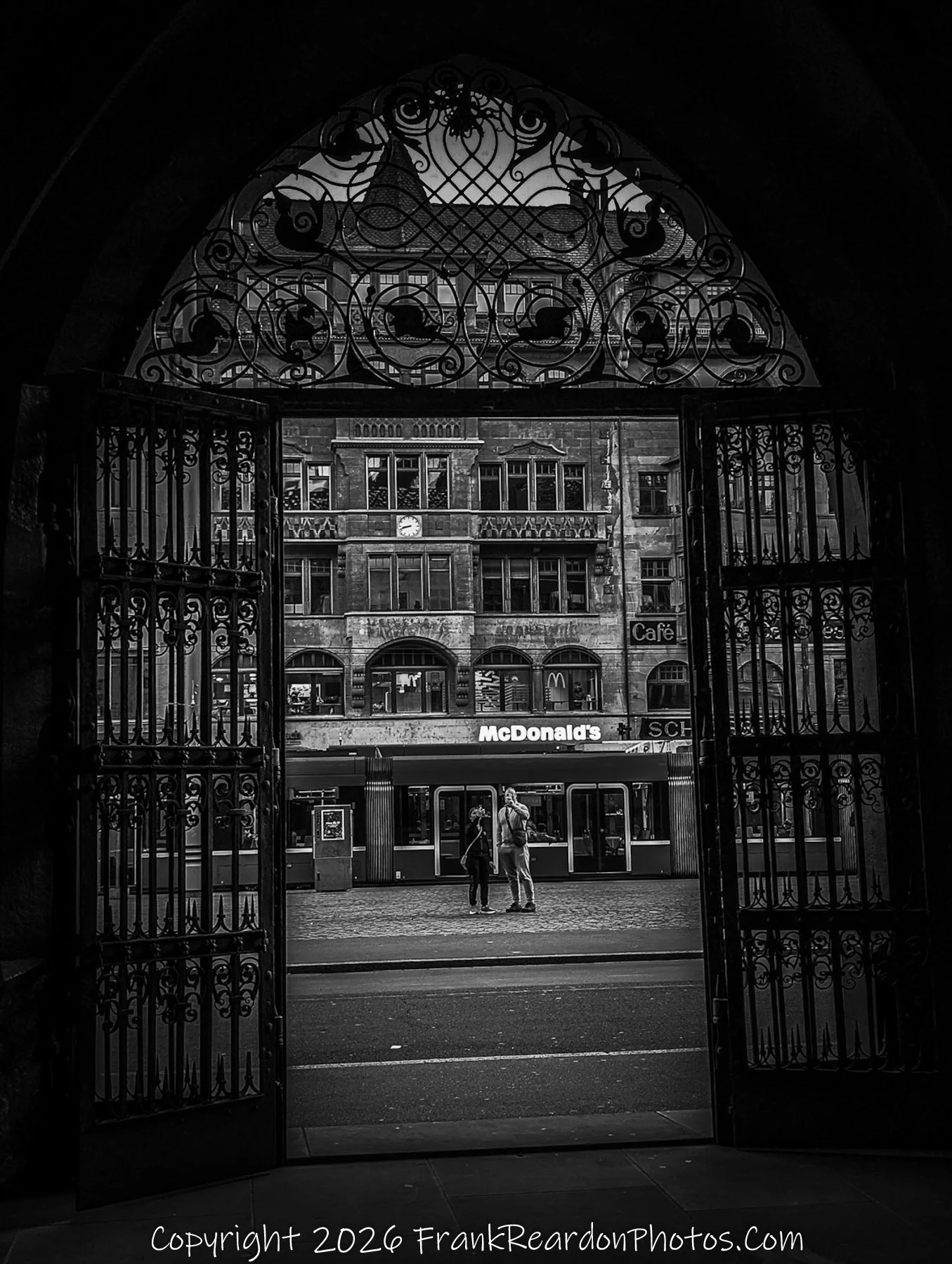 “Portal of Contrasts” The ornate iron gate frames a modern street scene, juxtaposing history and commerce in a single gaze.