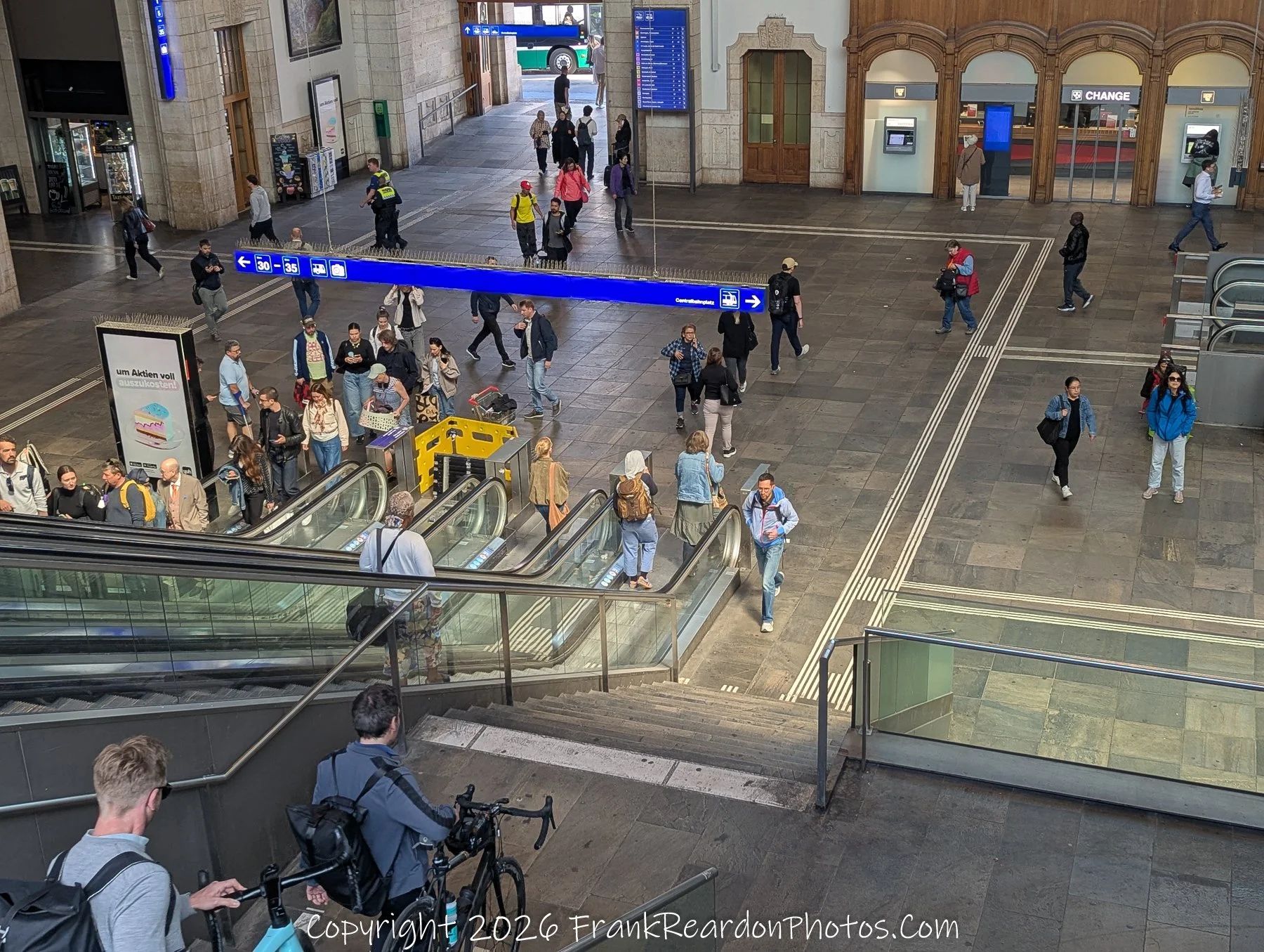 "Station Life" A top-down view of a bustling station concourse where people are walking toward escalators and signage, capturing the daily flow of travel.