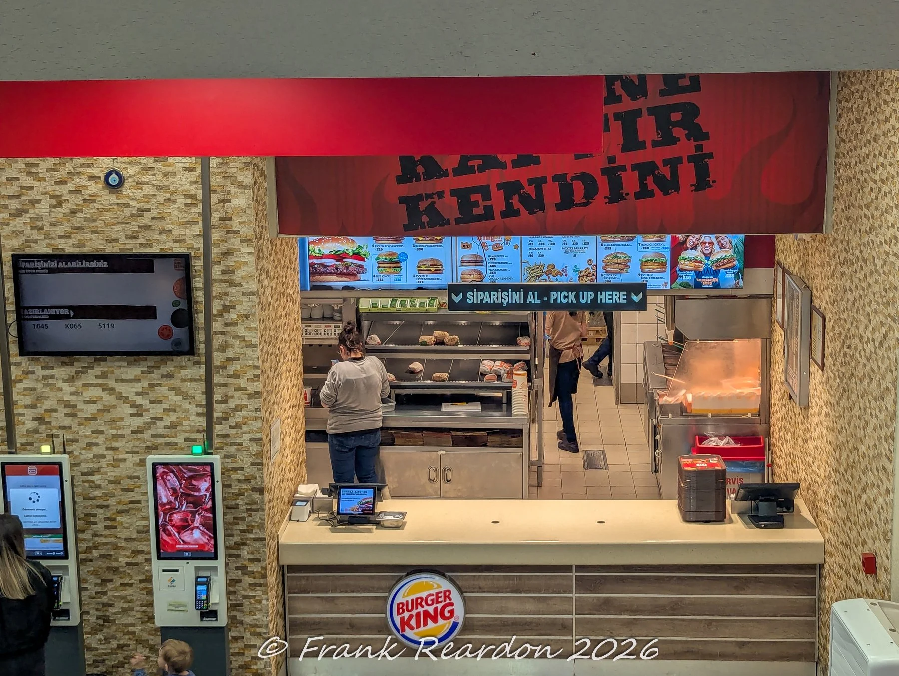 A high-angle interior shot looking down at a Burger King service counter with digital menu boards and staff members working behind the register.