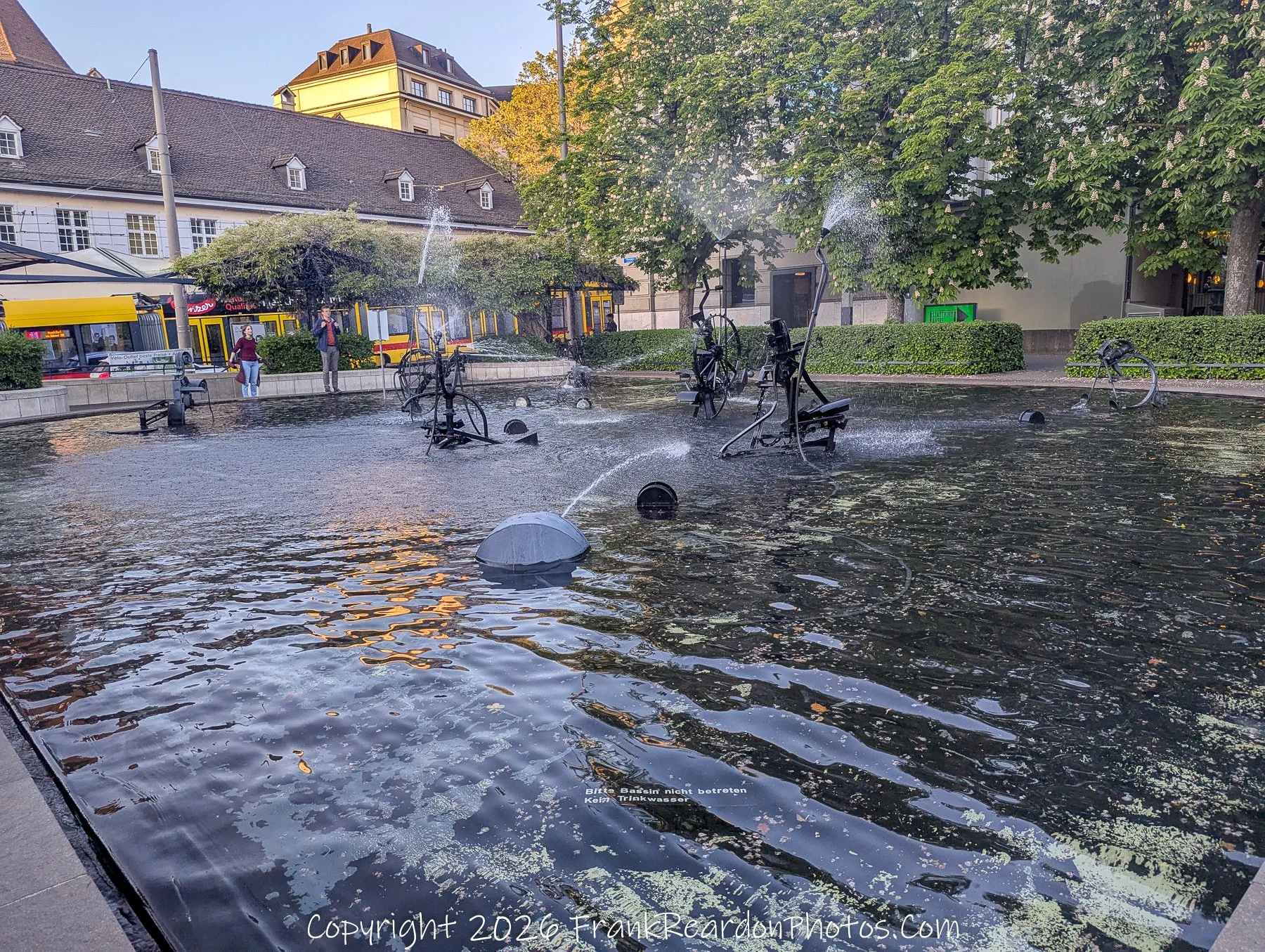 "Kinetic Bike Fountain" A public fountain featuring sculptural bicycle parts integrated into water jets, with a small sign warning not to enter the water.