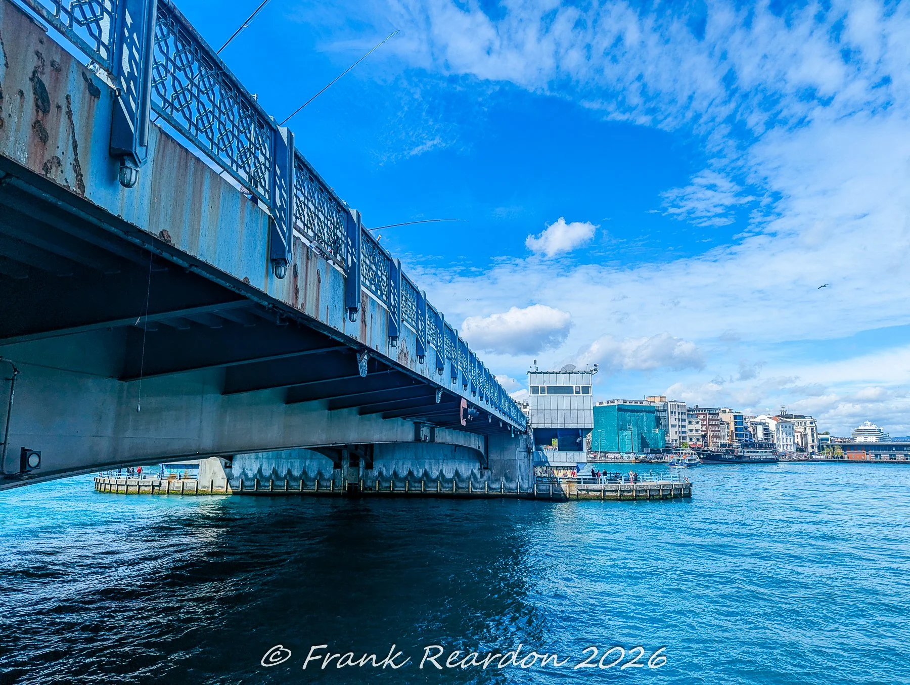 Looking up at the steel architecture and blue railings of the Galata Bridge spanning across the sparkling waters with city buildings in the background.