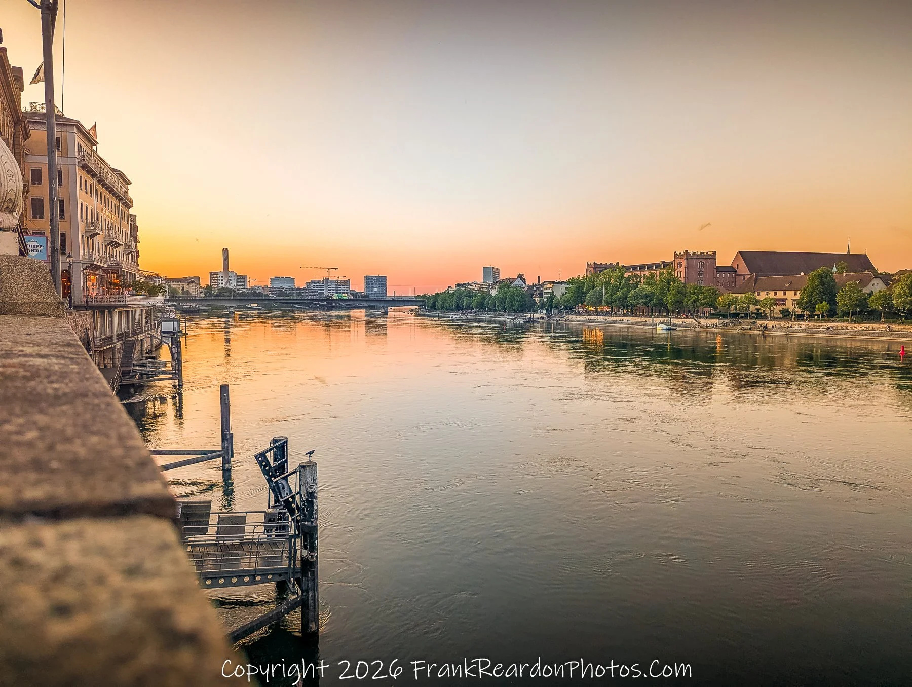 "Sunset Over the Rhine" A serene, wide-angle view of the Rhine River reflecting the vibrant orange hues of a sunset, looking out from a stone embankment towards the city buildings.