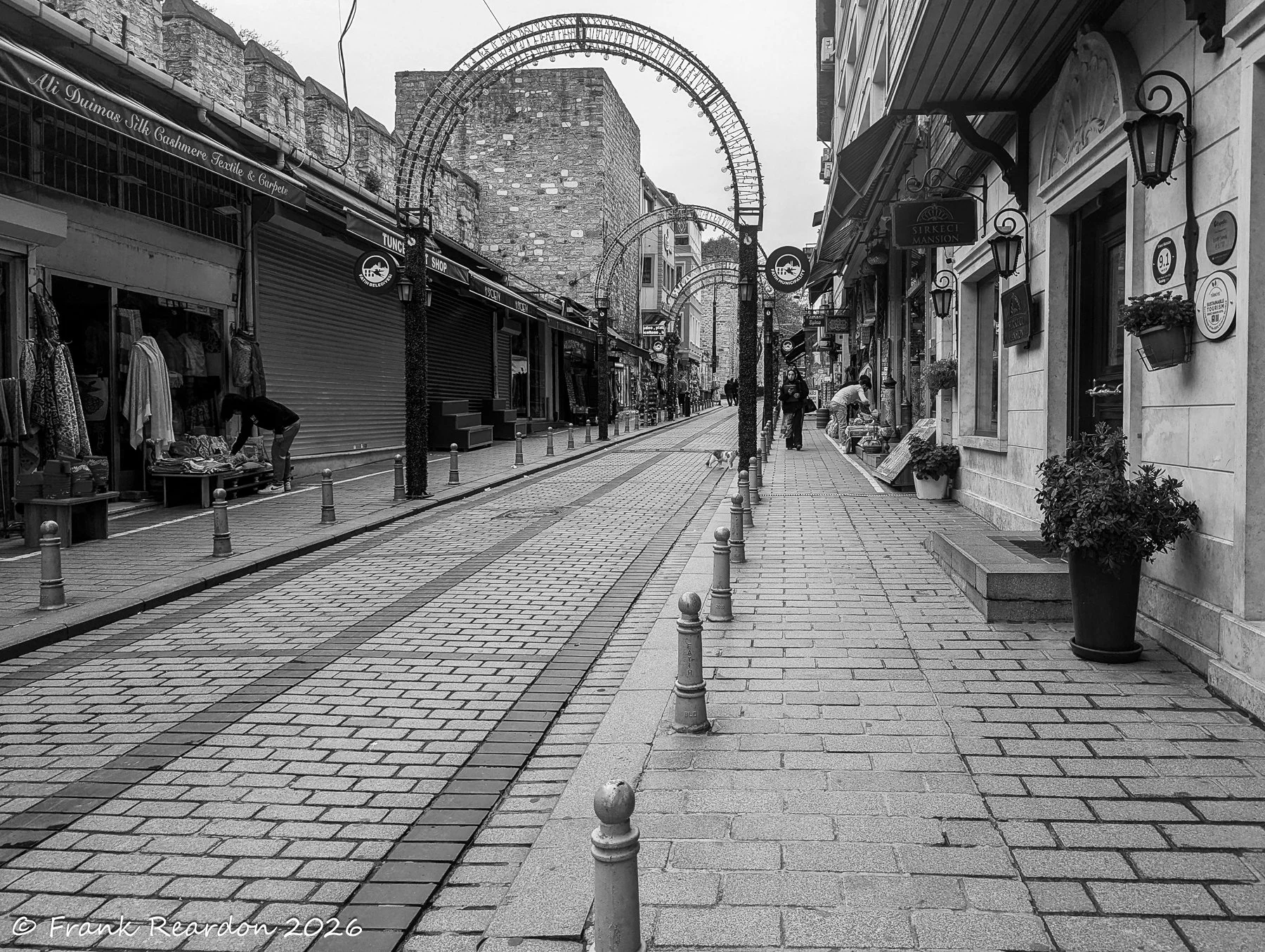 A moody monochrome perspective of an ancient Istanbul street, lined with historic buildings, shops, and overhead arches.