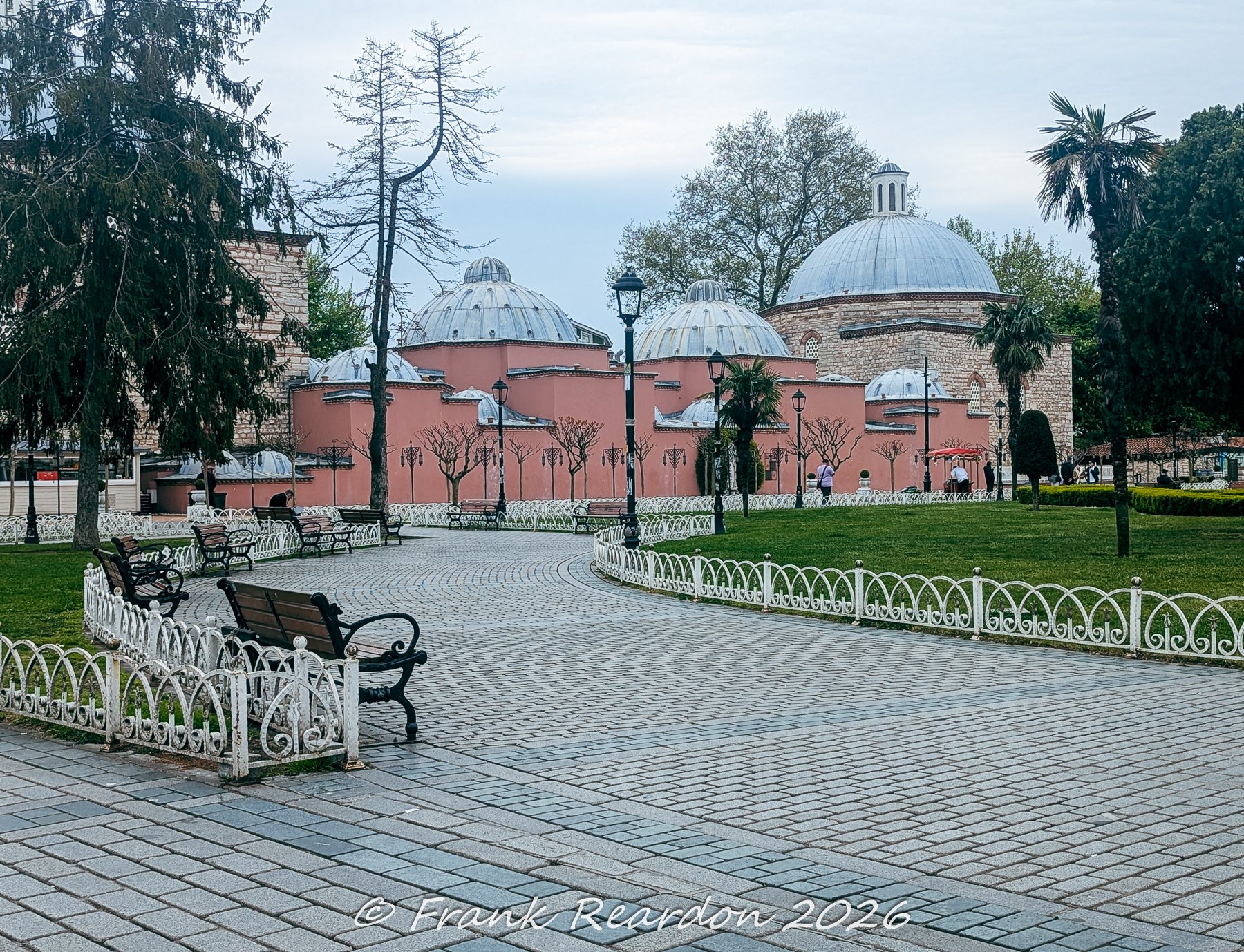 A peaceful, paved pathway flanked by benches and lush greenery leading toward classic domed architecture in Istanbul.