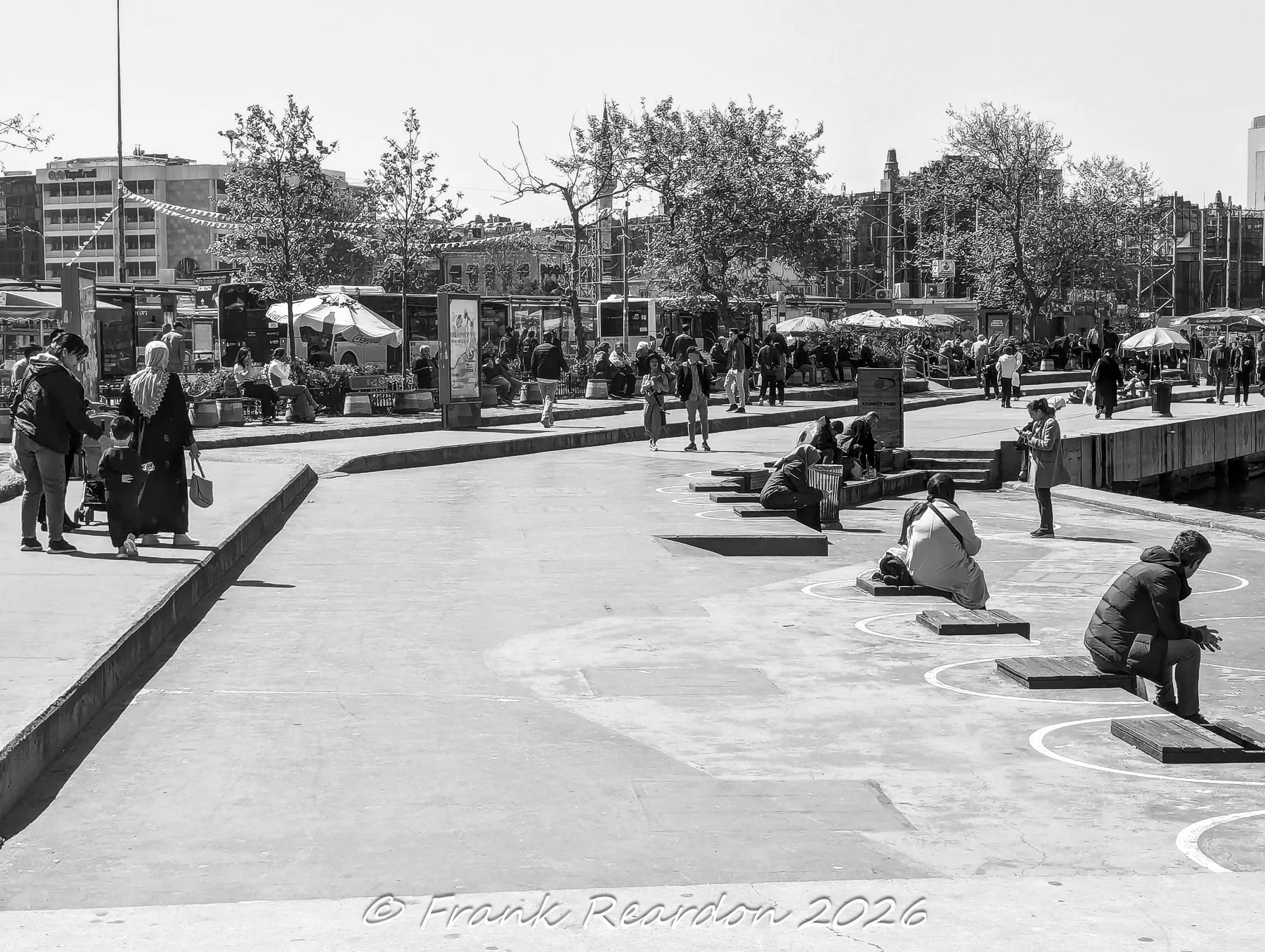 A black-and-white street scene capturing locals walking and resting on wooden block seating along a modern esplanade next to the water.