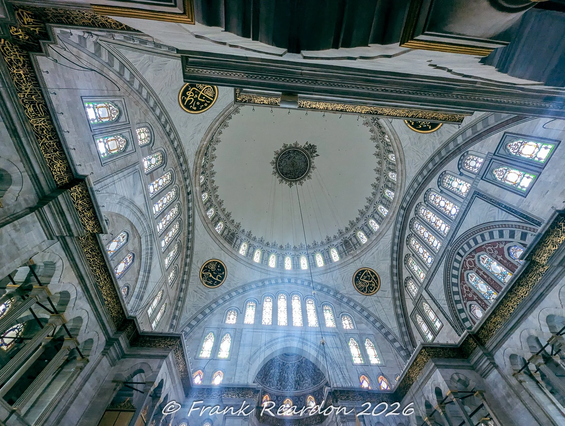 A stunning upward look at the soaring central dome, intricate stained-glass arched windows, and golden Arabic calligraphy inscribed along the interior walls of the Nuruosmaniye Mosque.