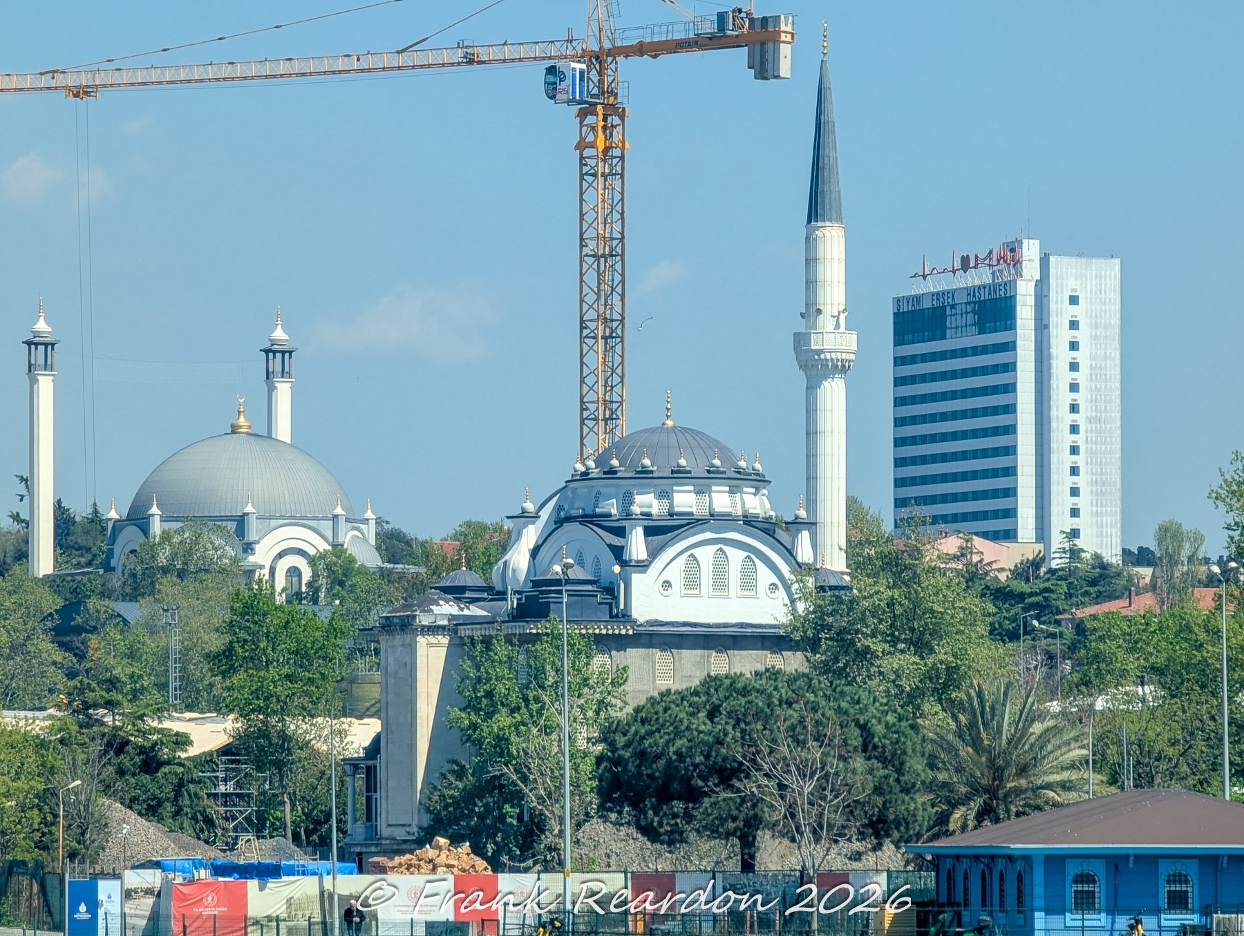 A view featuring a classic Ottoman-style mosque complex with domes and a slender minaret, framed by a construction crane and the modern Siyami Ersek Hospital building in the background.