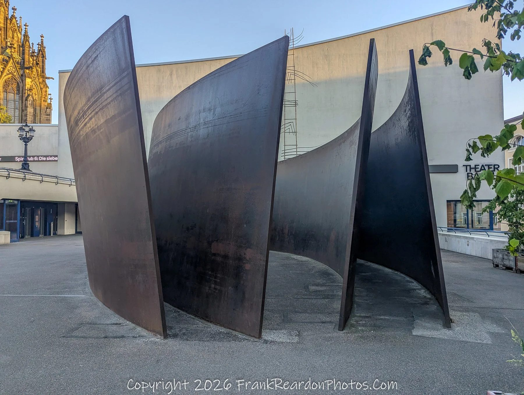 "Minimalist Steel Sculpture" A large, curved, weathered-steel sculpture installed in a plaza, positioned in front of a modern building facade with the sign "Theater Basel".