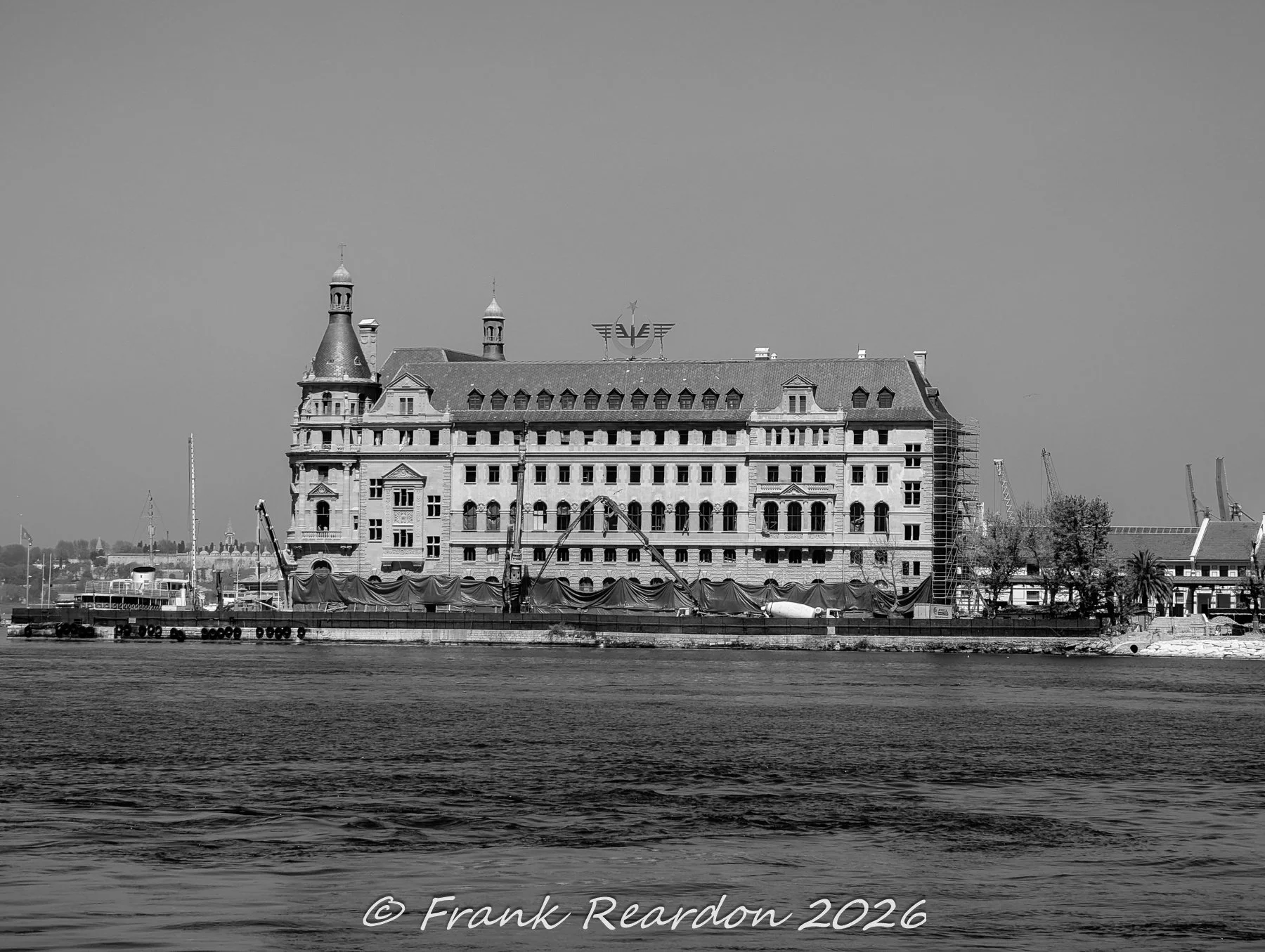 A wide black-and-white view of the grand, historic Haydarpaşa Railway Station building resting on the waterfront with scaffolding and protective wraps during its restoration project.