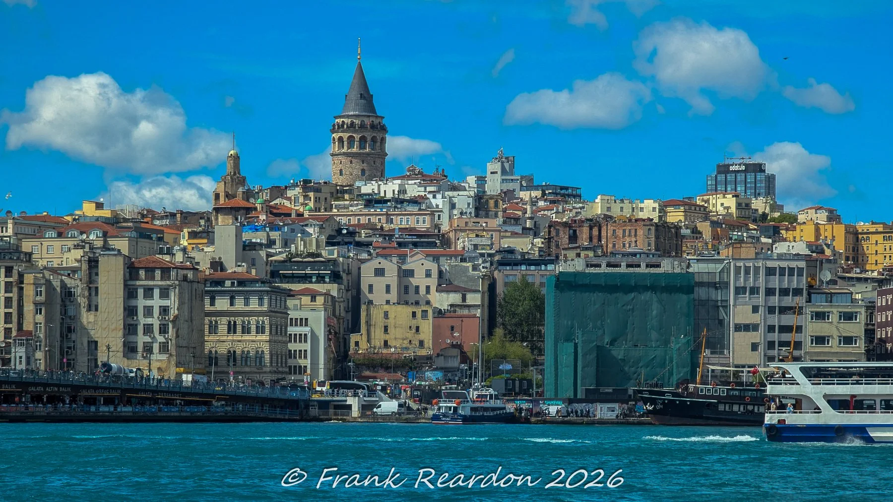 A scenic view across the water featuring the historic Galata Tower rising above the densely built hillside, classic rooftops, and the bustling waterfront of Istanbul.