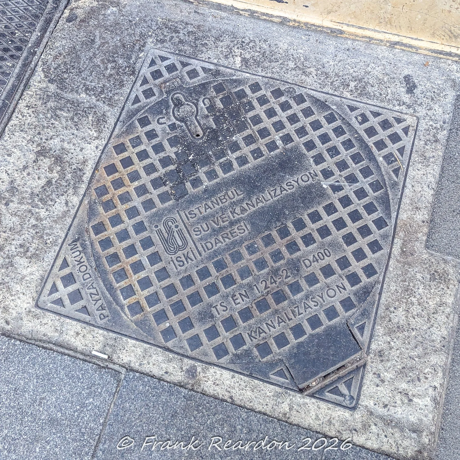 A close-up view of a metal utility cover embedded in the pavement bearing the emblem and text for Istanbul Su ve Kanalizasyon İdaresi (İSKİ).