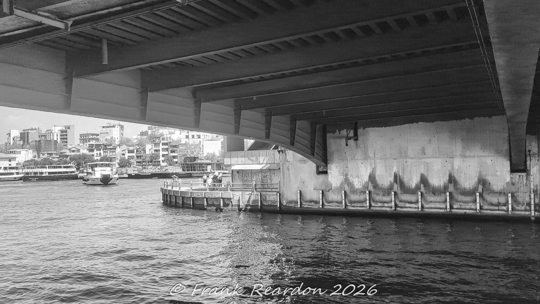 A black-and-white perspective looking out from under the heavy concrete and steel framework of the Galata Bridge toward the waters and buildings of the Golden Horn.