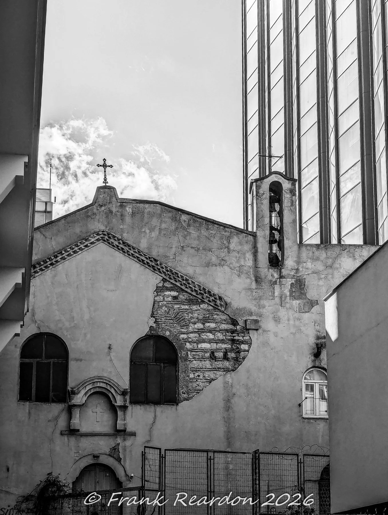 A black-and-white architectural shot showing the weathered stone and arched windows of a historic church structure nestled right next to a modern glass high-rise.