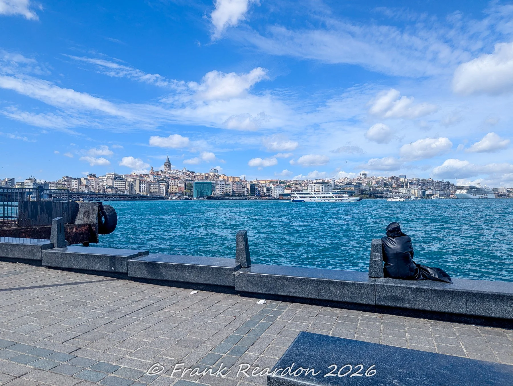 A person sitting on the stone seawall along the bustling waterfront, looking out across the blue waters toward the Galata Tower and the historic city skyline.