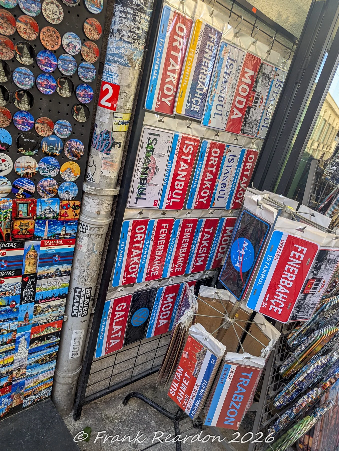 A vibrant storefront display showcasing a large collection of decorative metal neighborhood signs (such as Ortaköy, Fenerbahçe, and Moda) alongside colorful souvenir magnets.