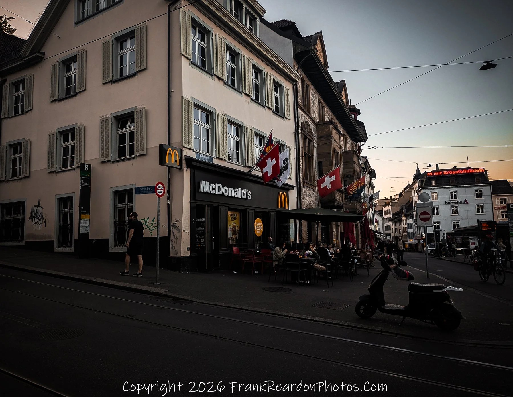 "McDonald’s at Dusk" A McDonald's restaurant situated on a city corner, decorated with Swiss flags, featuring outdoor seating and a parked scooter.