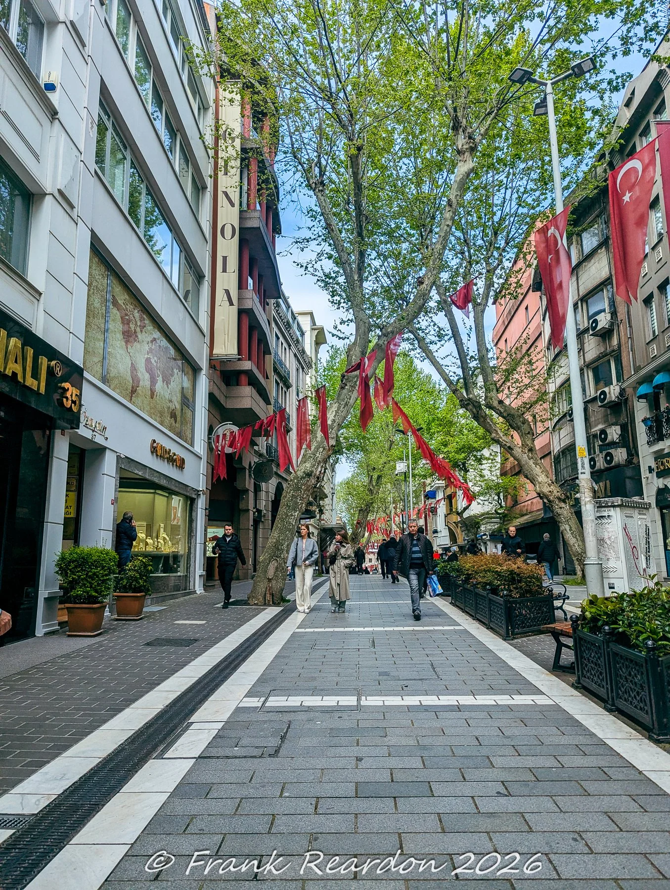 A bright daytime view looking down a paved shopping and pedestrian street lined with historic buildings, trees, and hanging flags.