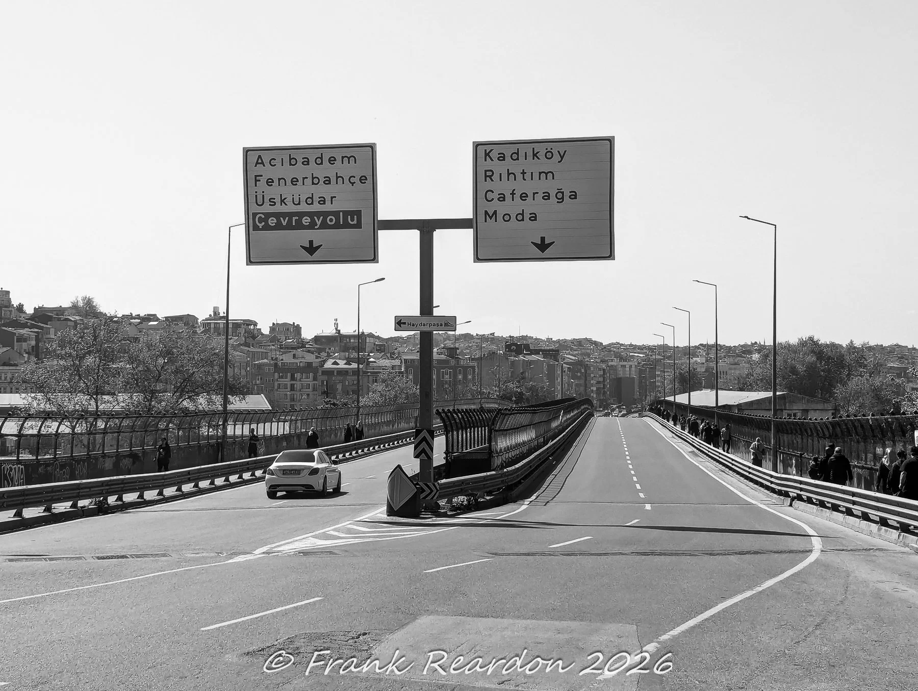 A black-and-white shot looking down a paved roadway toward overhead directional signs pointing toward Acıbadem, Fenerbahçe, Üsküdar, Kadıköy, and Moda.